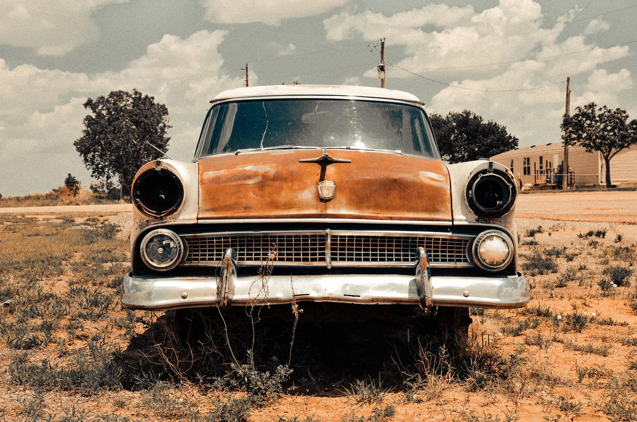 An old, rusty vintage car missing its headlights, parked on a dry, grassy area with a partly cloudy sky in the background.