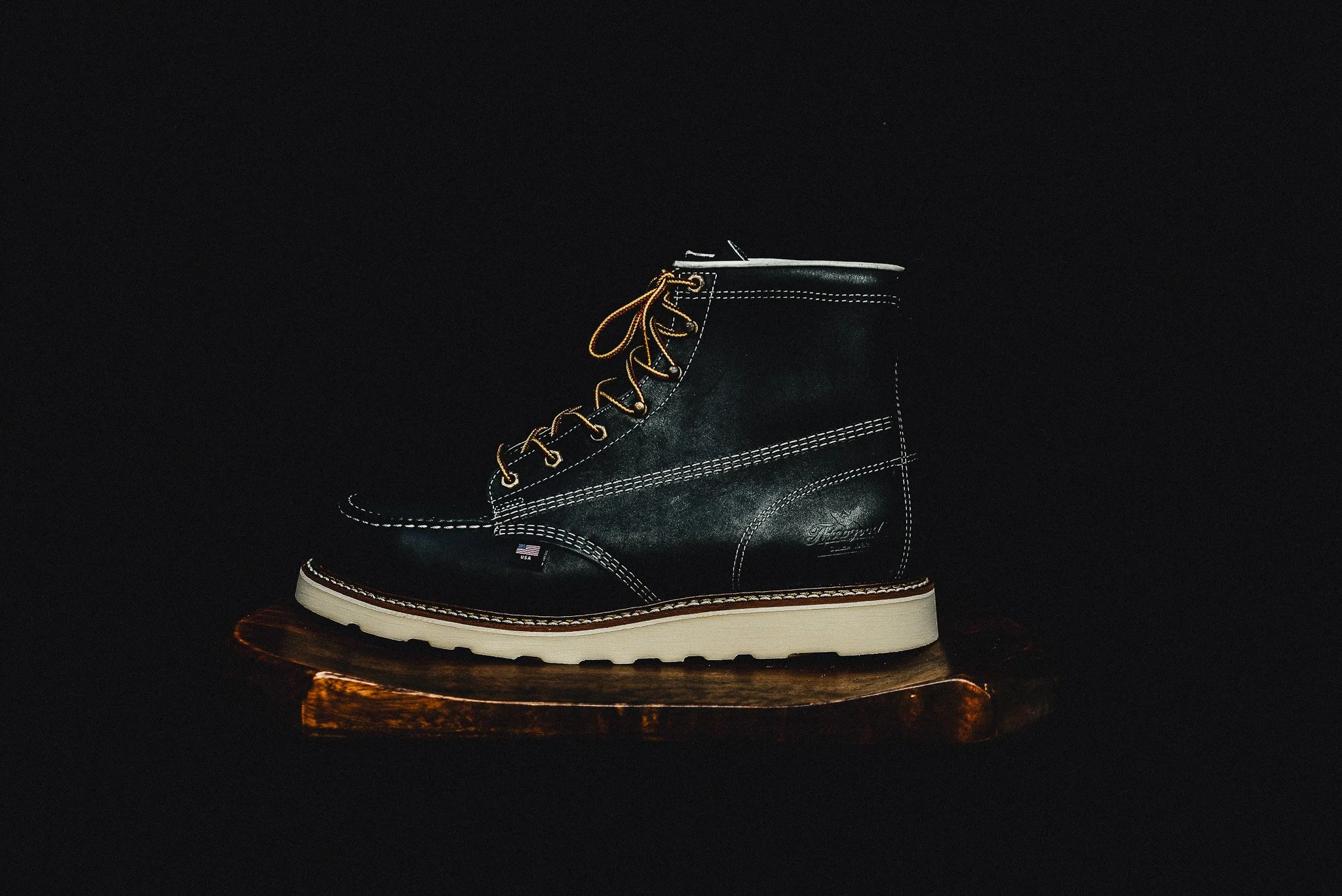 A black leather boot with white stitching and yellow laces, displayed on a wooden platform against a dark background.