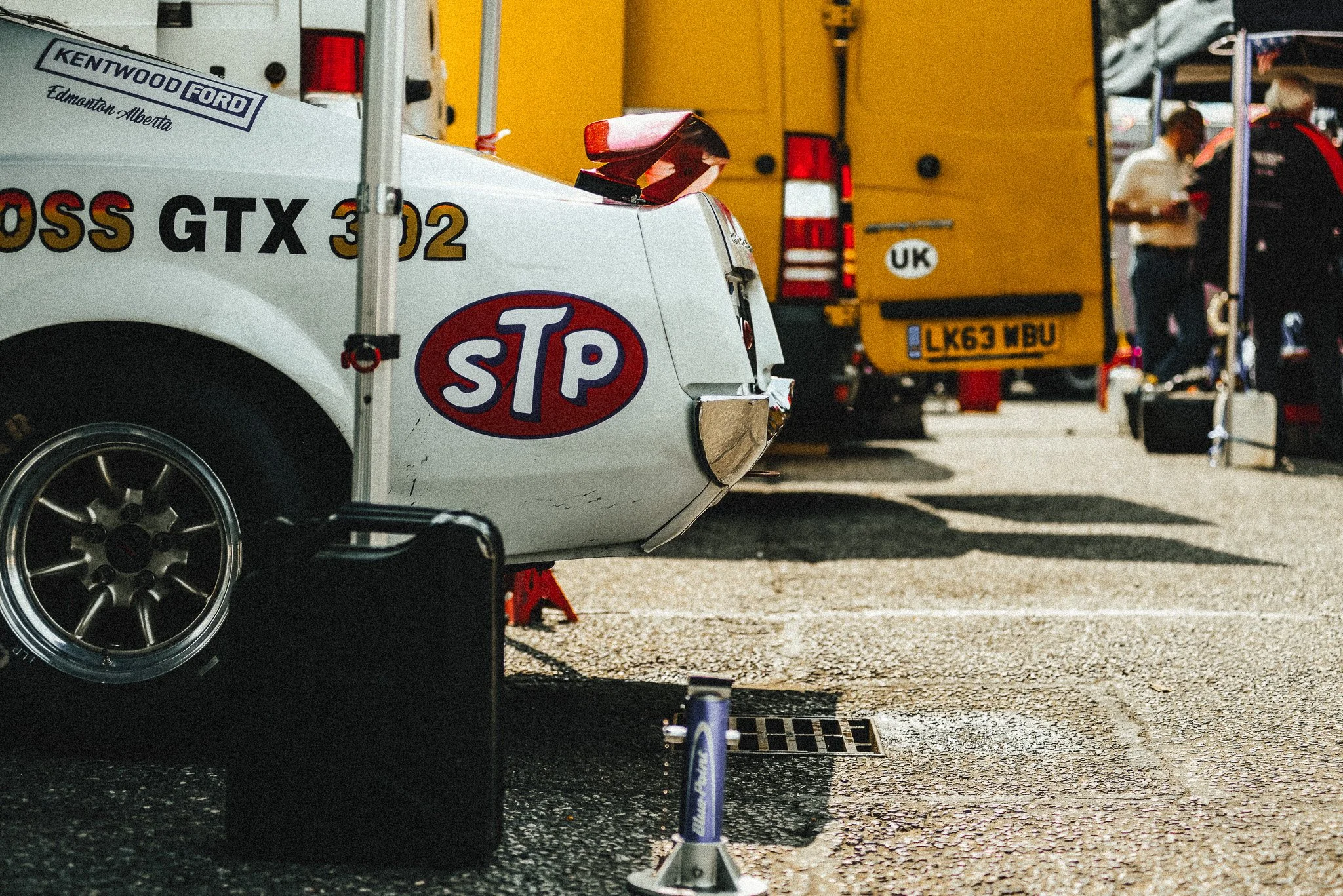 Part of a vintage race car with a STP logo, yellow trucks in the background, and a person working at a racing setup in a paddock area.