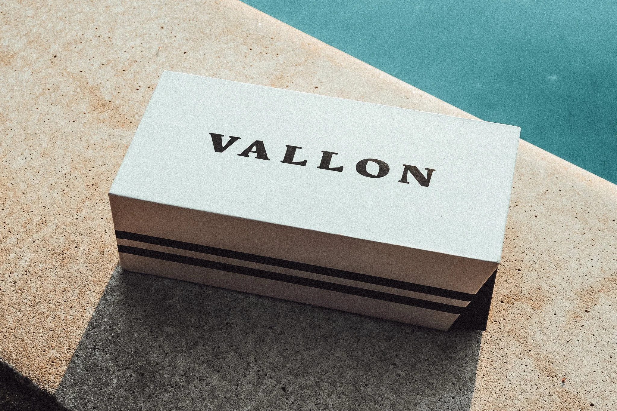 A white box with the word 'VALLON' in black, placed on a concrete surface near a swimming pool.