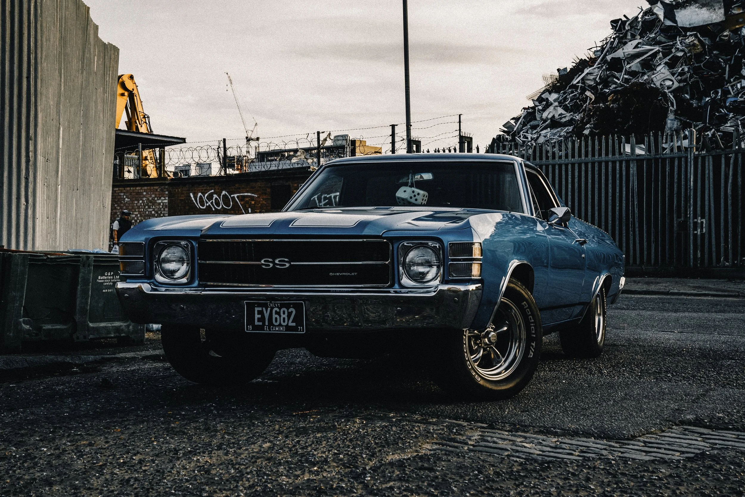 A vintage blue Chevrolet Chevelle SS muscle car parked in an industrial area with scrap metal piles and graffiti in the background.