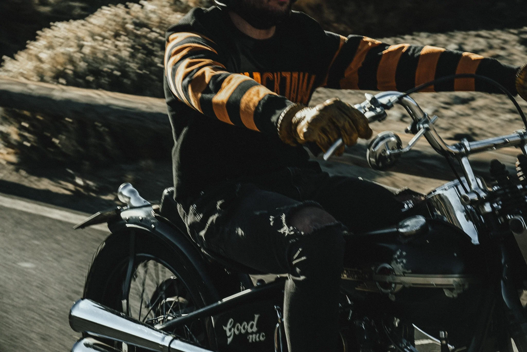 Person riding a black motorcycle on a road, wearing orange and black striped long-sleeve shirt, black gloves, and ripped black jeans.