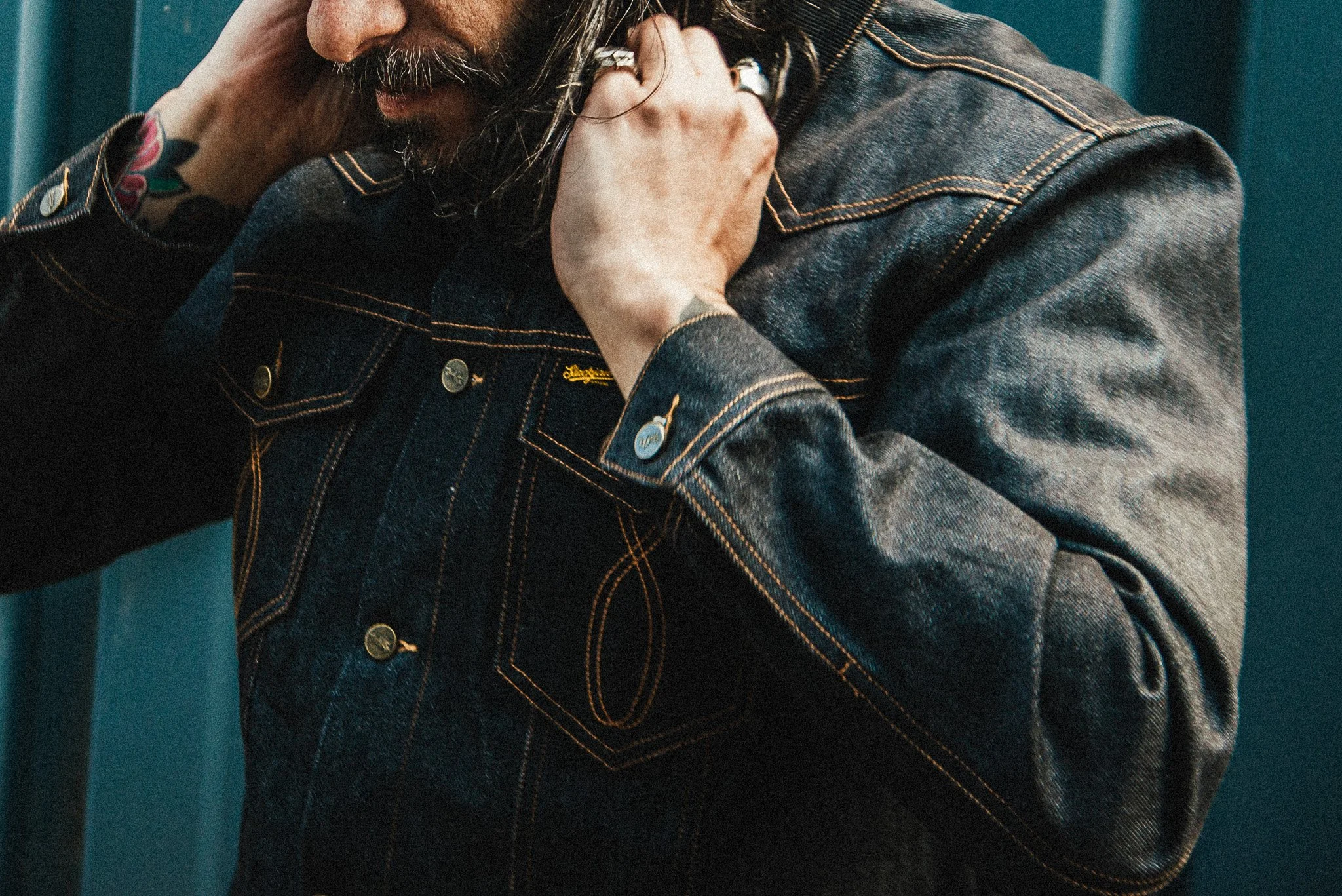 A man wearing a dark denim jacket with yellow stitching, adjusting his collar near his face, with a floral tattoo on his left wrist and rings on his fingers.