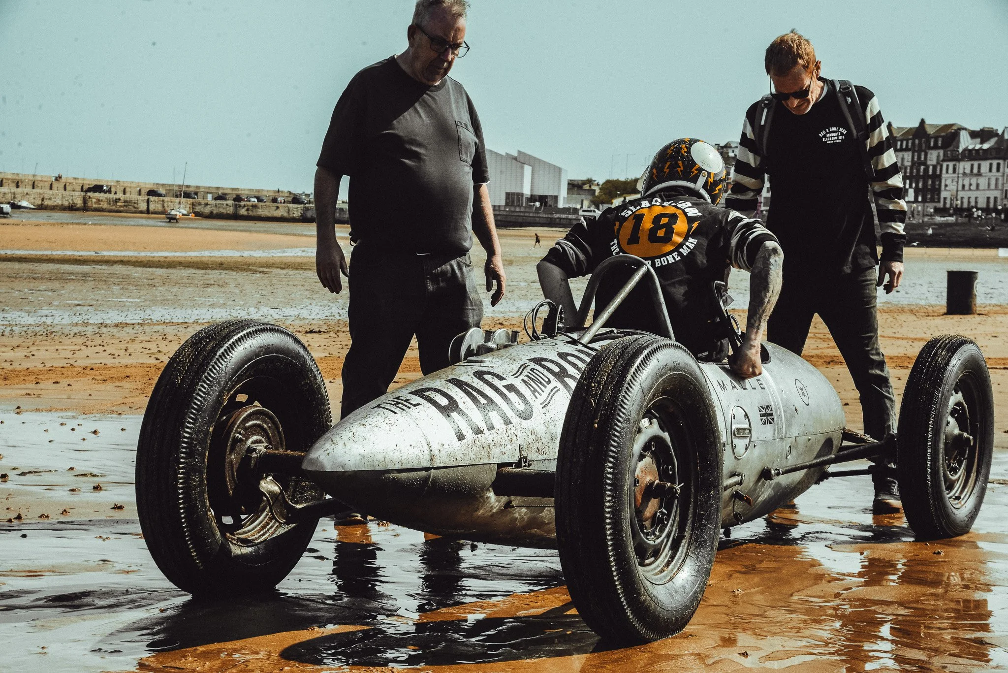 Three men and a vintage race car on a beach, with buildings and a harbor in the background.