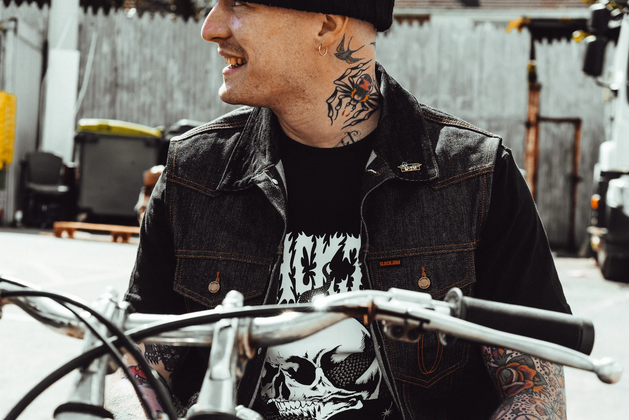 Close-up of a smiling man with tattoos on neck and arms, wearing a black denim vest and black T-shirt with a skull design, sitting on a bicycle in an outdoor setting.