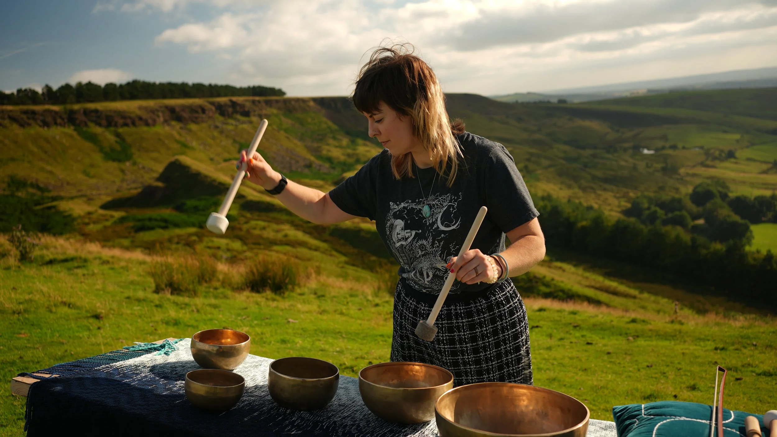 Clare is poised with a mallet mid-air about to dong one of several Himalayan singing bowls placed on a table. The backdrop is the beautiful green sweeping view of the Peak District.