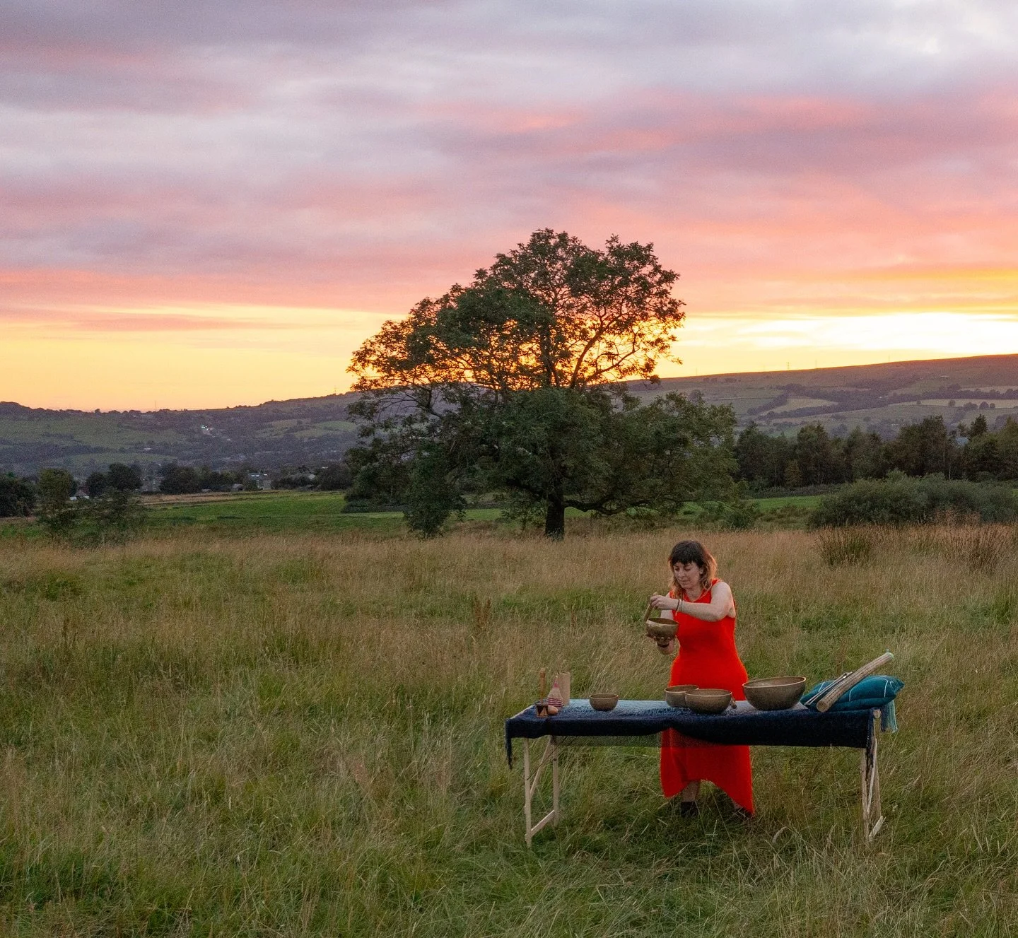 Sound therapist playing singing bowls outdoors in nature at sunset, offering a sound bath therapy session for relaxation and stress relief.