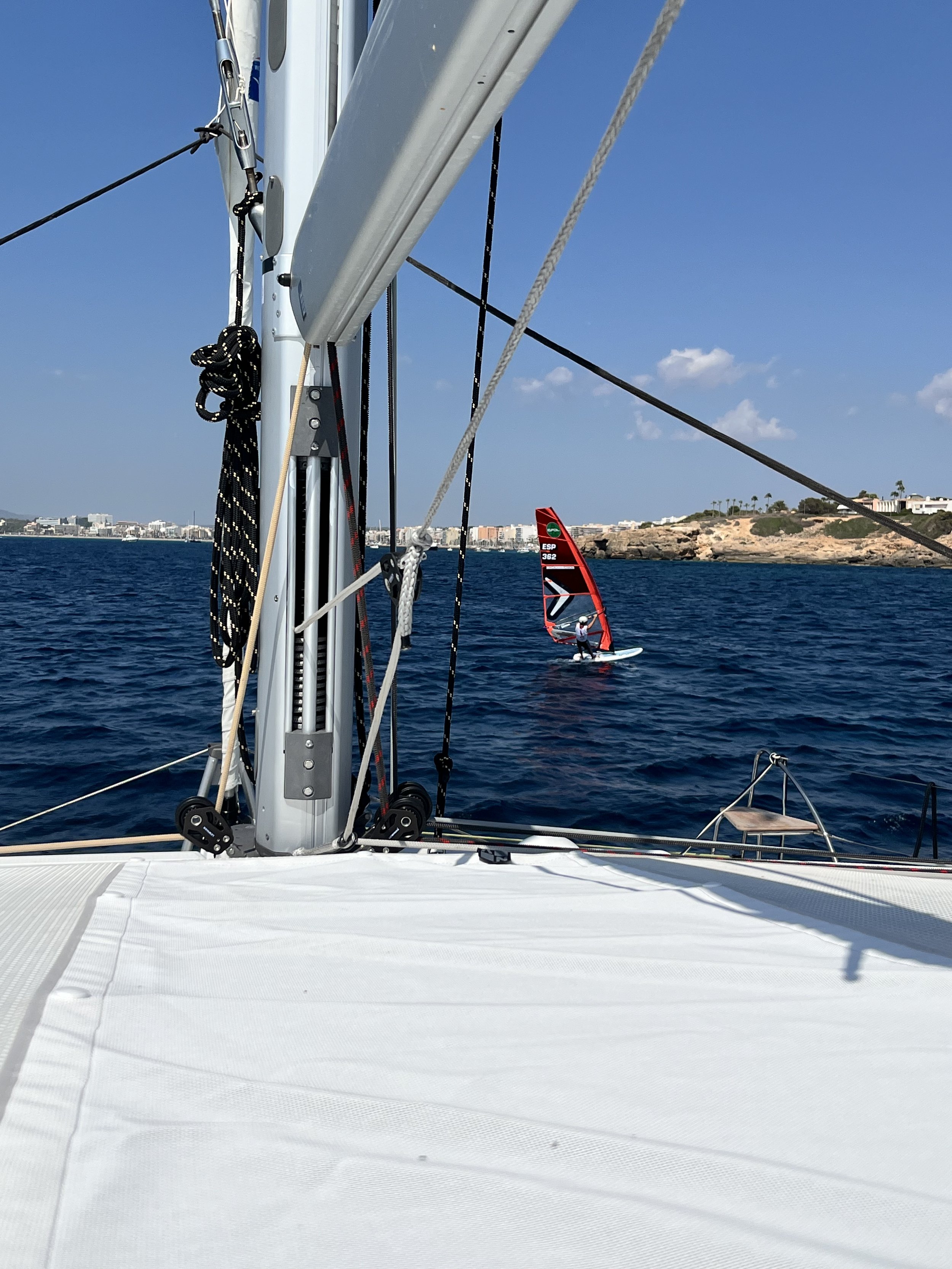 View from the deck of a sailboat showing the sailboat's mast and rigging, a windsurfer with a red sail in the water, and a distant shoreline with buildings, rocks, and some palm trees under a clear blue sky.