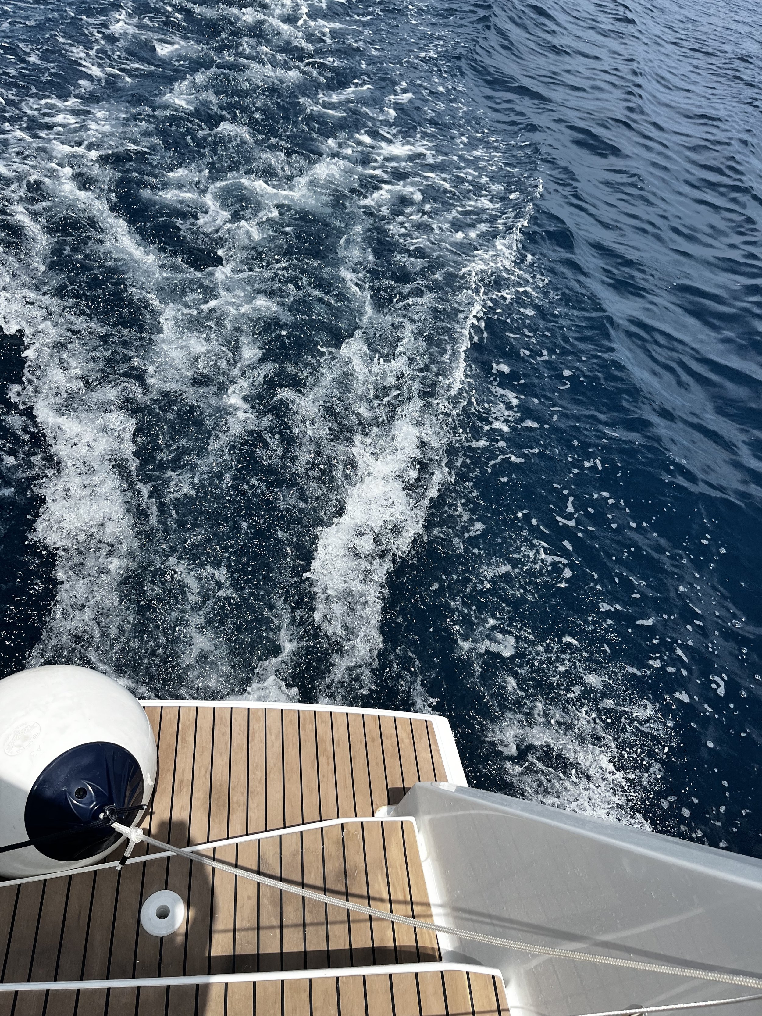 View of a boat's stern with a wooden deck, a white fender, and a rope, leaving a wake in the blue ocean water.