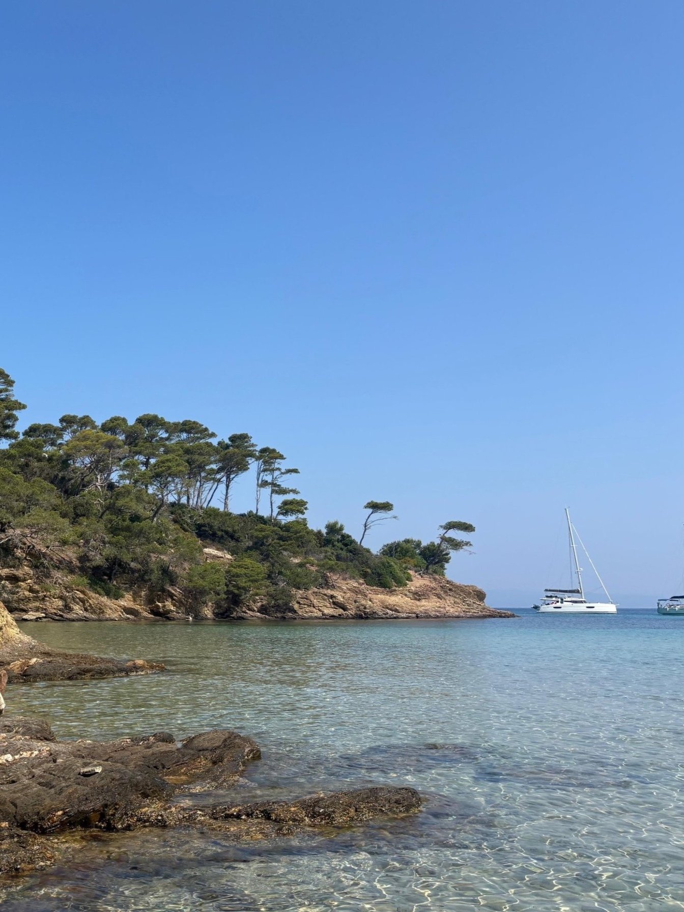 A scenic view of a rocky shoreline with clear water, a small forested hill with sparse trees, and sailboats anchored near the coast under a bright blue sky.