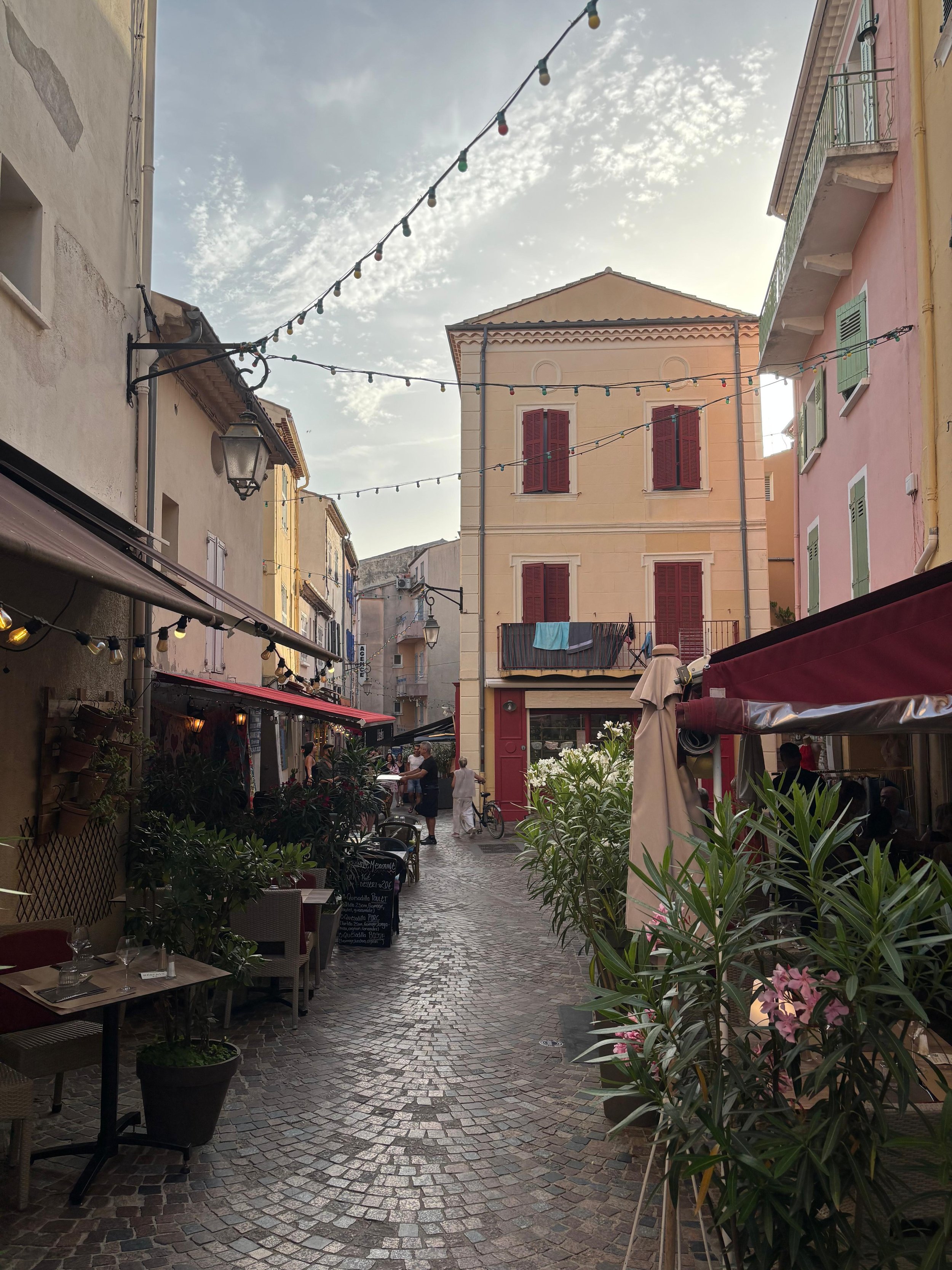A narrow cobblestone street in a European town lined with outdoor cafes, small shops, and pastel-colored buildings, with string lights overhead and a few people walking or sitting outside.