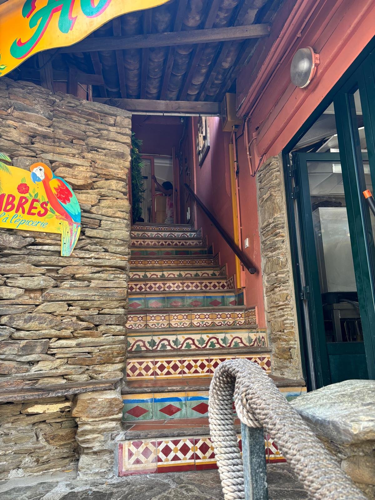 Colorful tiled stairs leading up to a restaurant or shop with a sign that says 'Tampires' and features a parrot. The stairs are decorated with intricate patterns, and there is a stone wall on the left. A person is partially visible at the top of the
