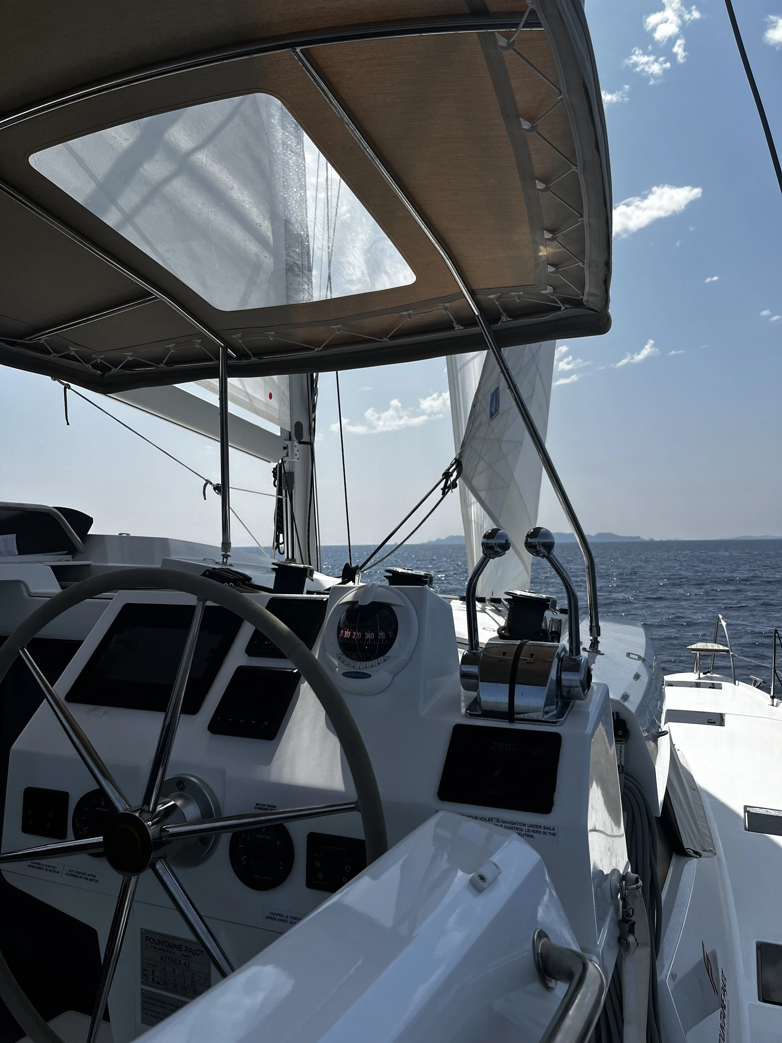 View of the interior of a sailboat's cockpit with steering wheel, navigational instruments, and a canopy, looking out over the ocean with blue sky and clouds.
