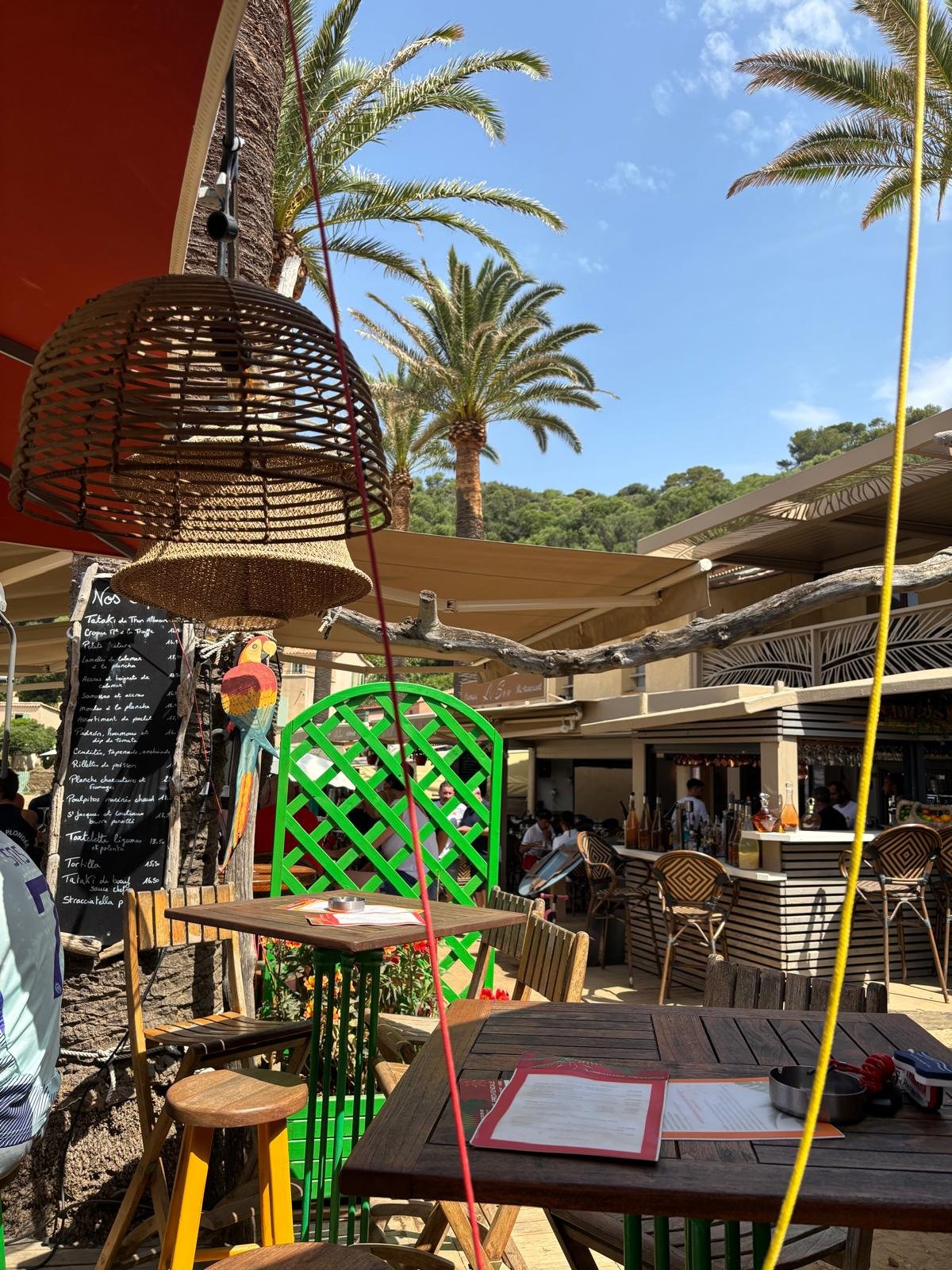 Outdoor restaurant with wooden tables and chairs, menu on the table, palm trees, blue sky, and a bar area in the background.