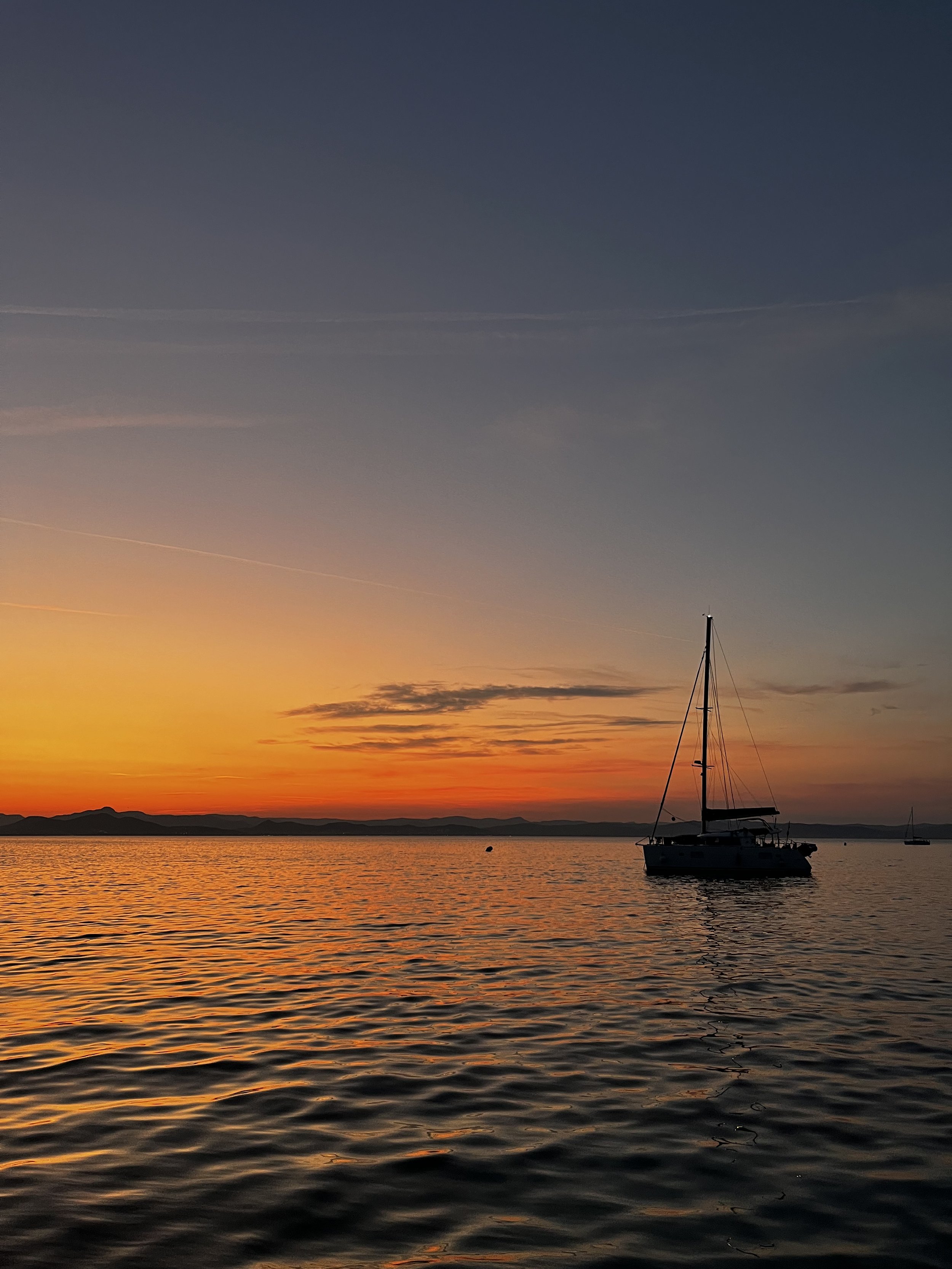 Sailboat anchored on calm water during a colorful sunset with a distant mountain range.