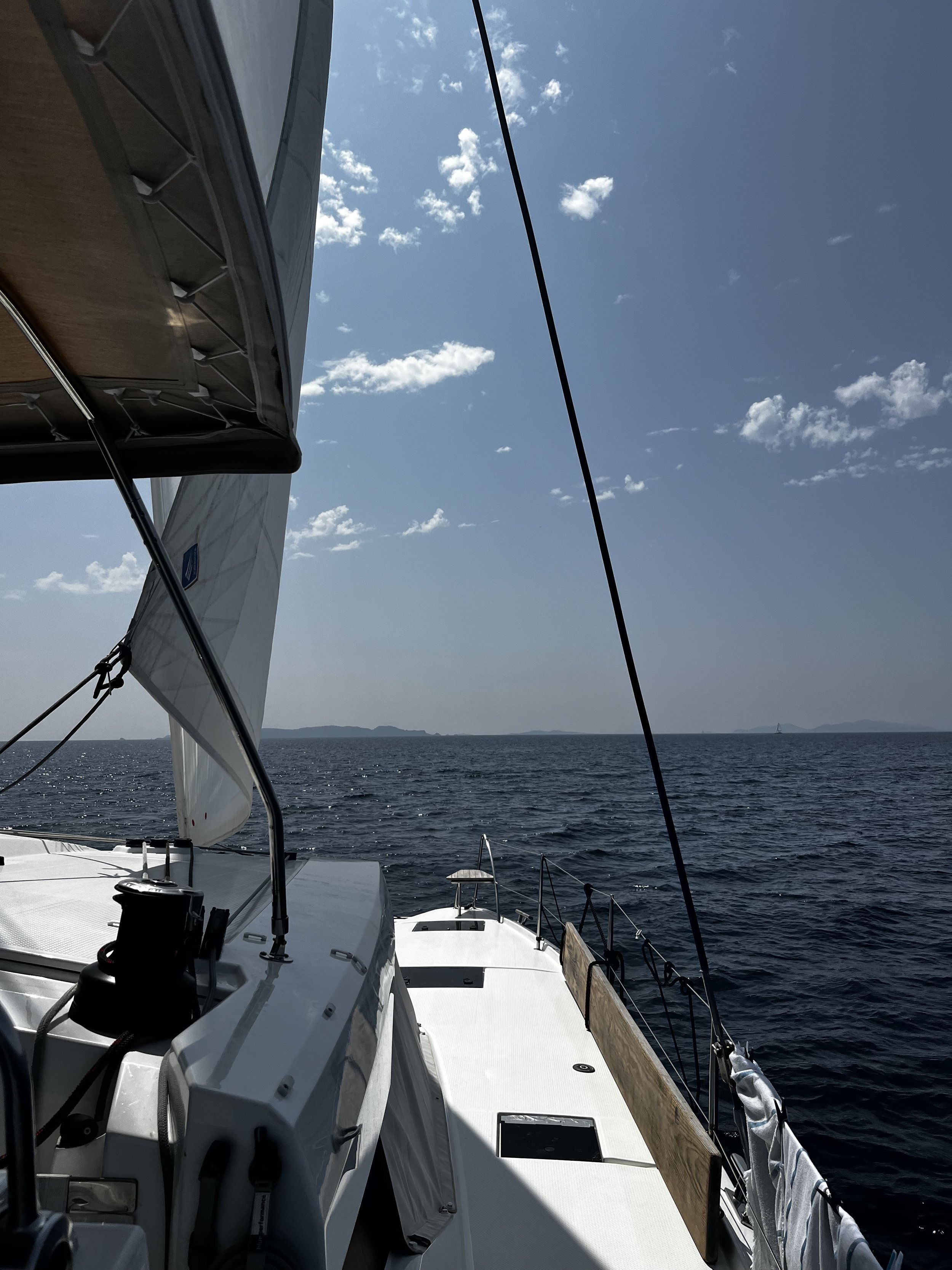 View from a sailboat on the open water, with the boat's deck and sail visible, under a bright blue sky with scattered clouds.