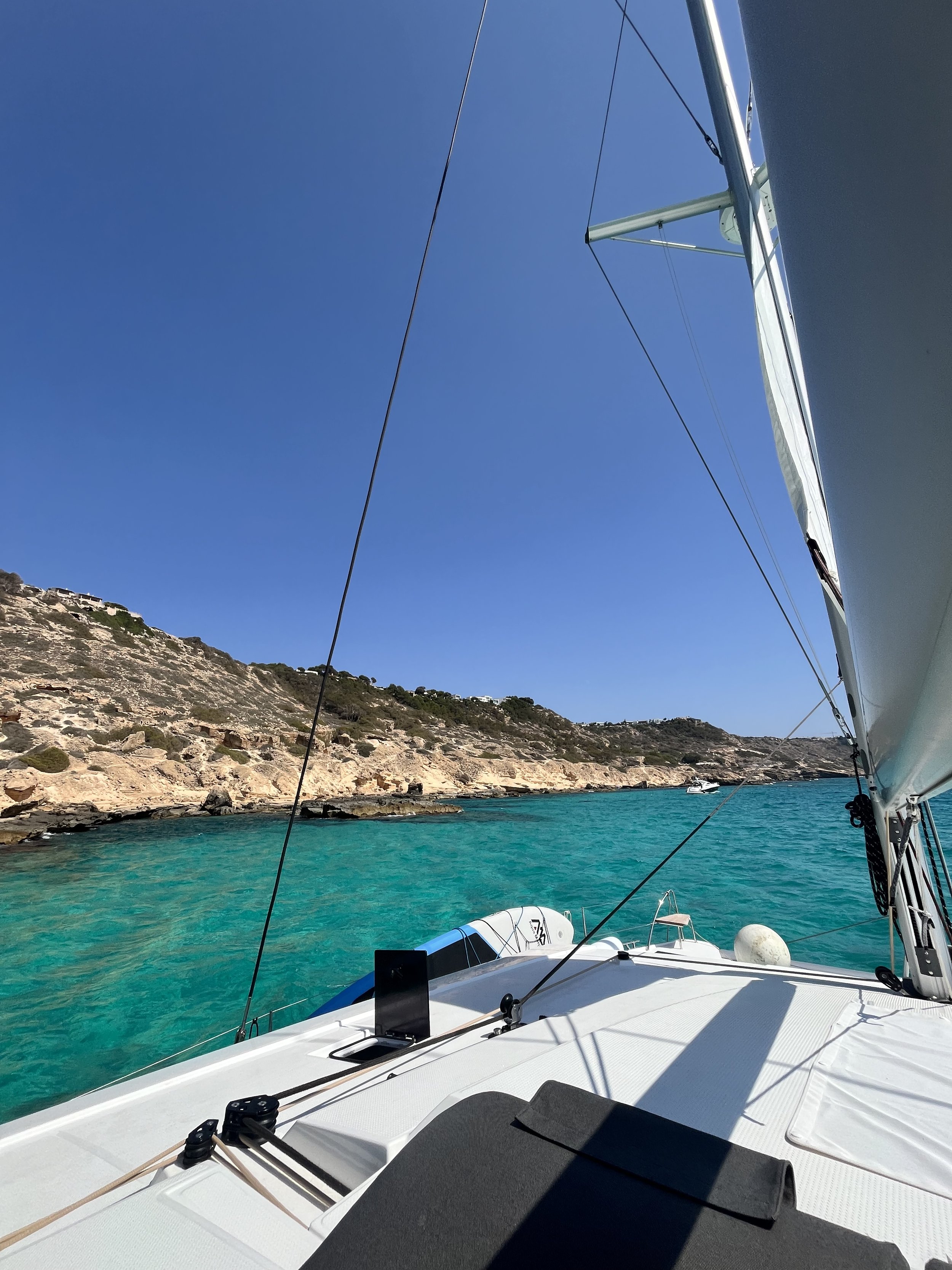 View from a sailboat looking towards a rocky coastline with clear blue water under a bright blue sky.
