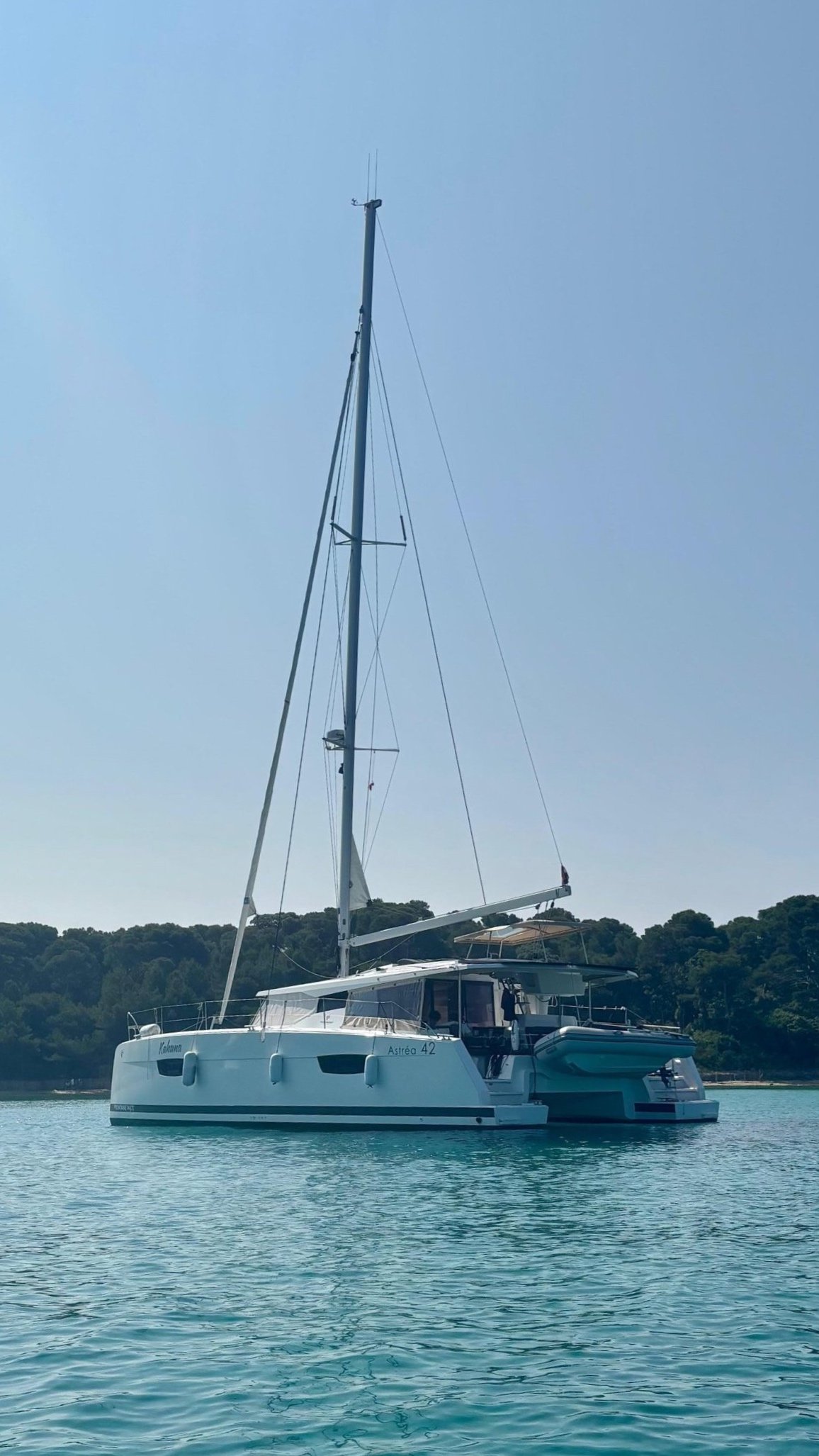 A white sailing yacht named Kahuna floating on calm water with a wooded shoreline in the background.