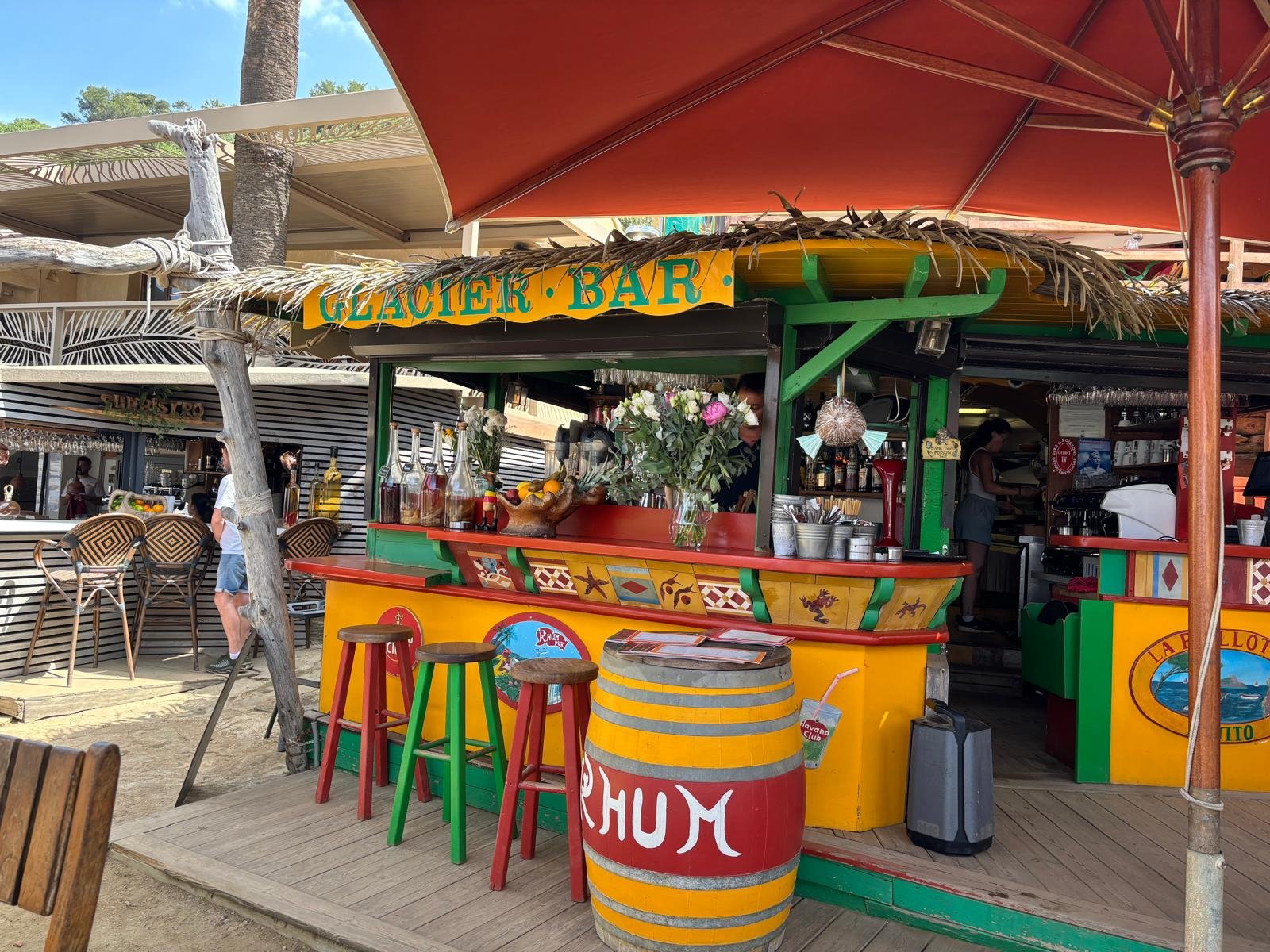 Colorful outdoor bar named 'Glacier Bar' with a yellow and red barrel-shaped table, three stools, flowers, and bar supplies, under a tan and red patio umbrella.