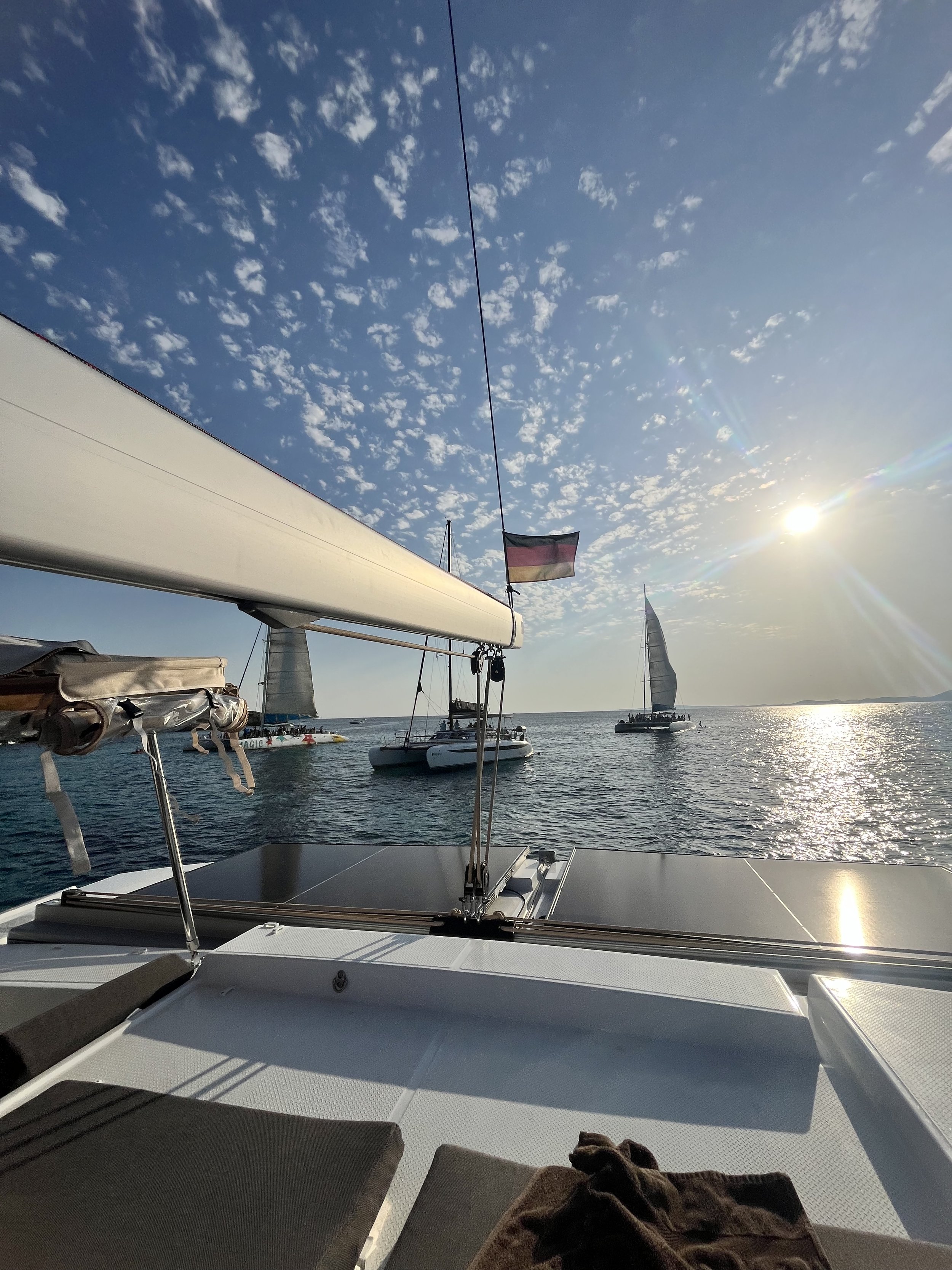View from a sailboat's deck looking out over the water with two other sailboats in the distance under a partly cloudy sky with the sun shining.