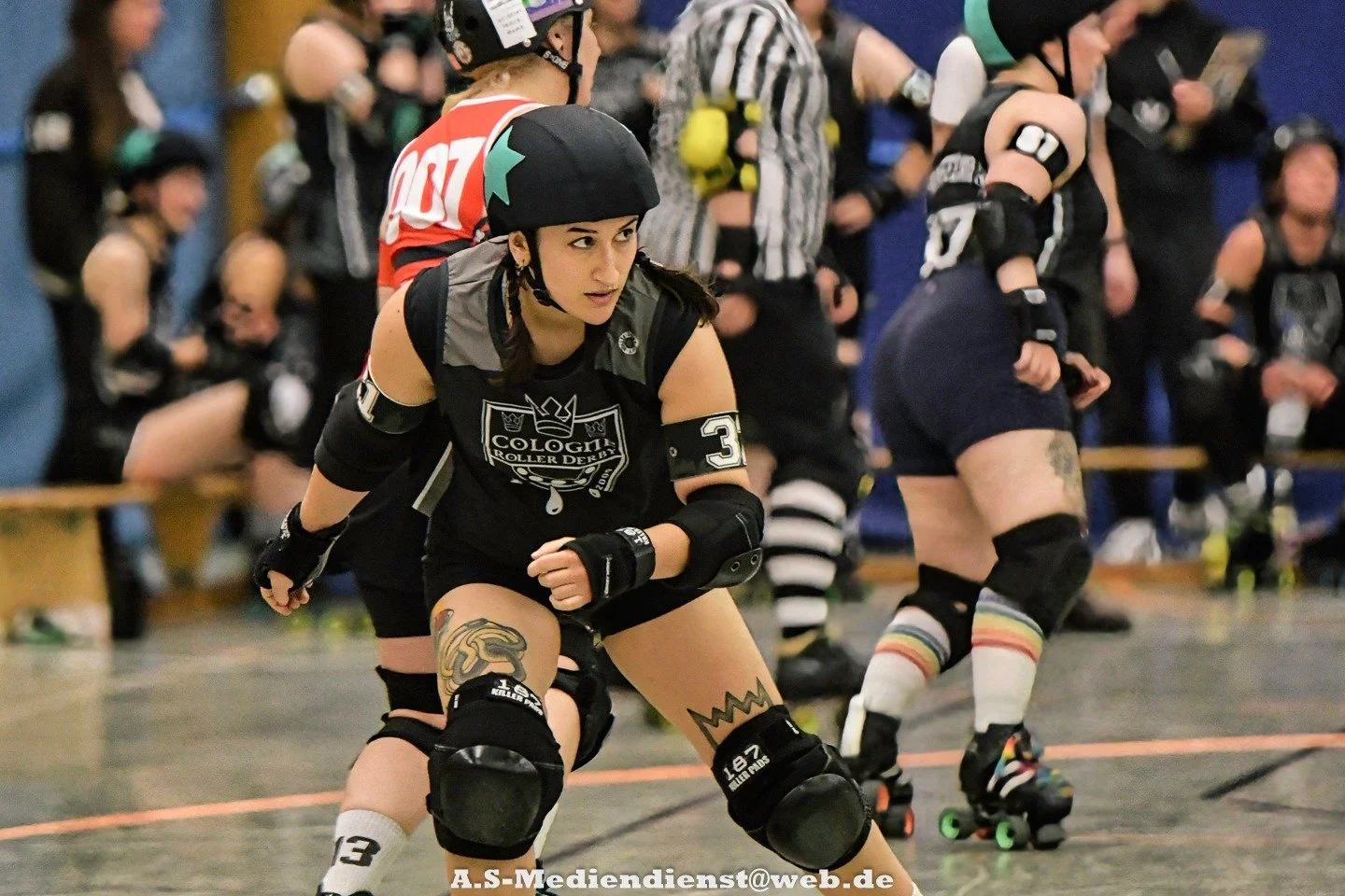 Roller Derby-Spielerin in Schutzausrüstung in Position, hinter ihr mehrere Mitspieler und Zuschauer