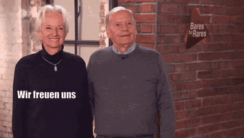 Ein älteres Paar steht lächelnd vor einer roten Backsteinwand im Studio, mit einem Schild im Hintergrund, das 'Bares für Rares' zeigt, und der Text 'Wir freuen uns' im Bild.