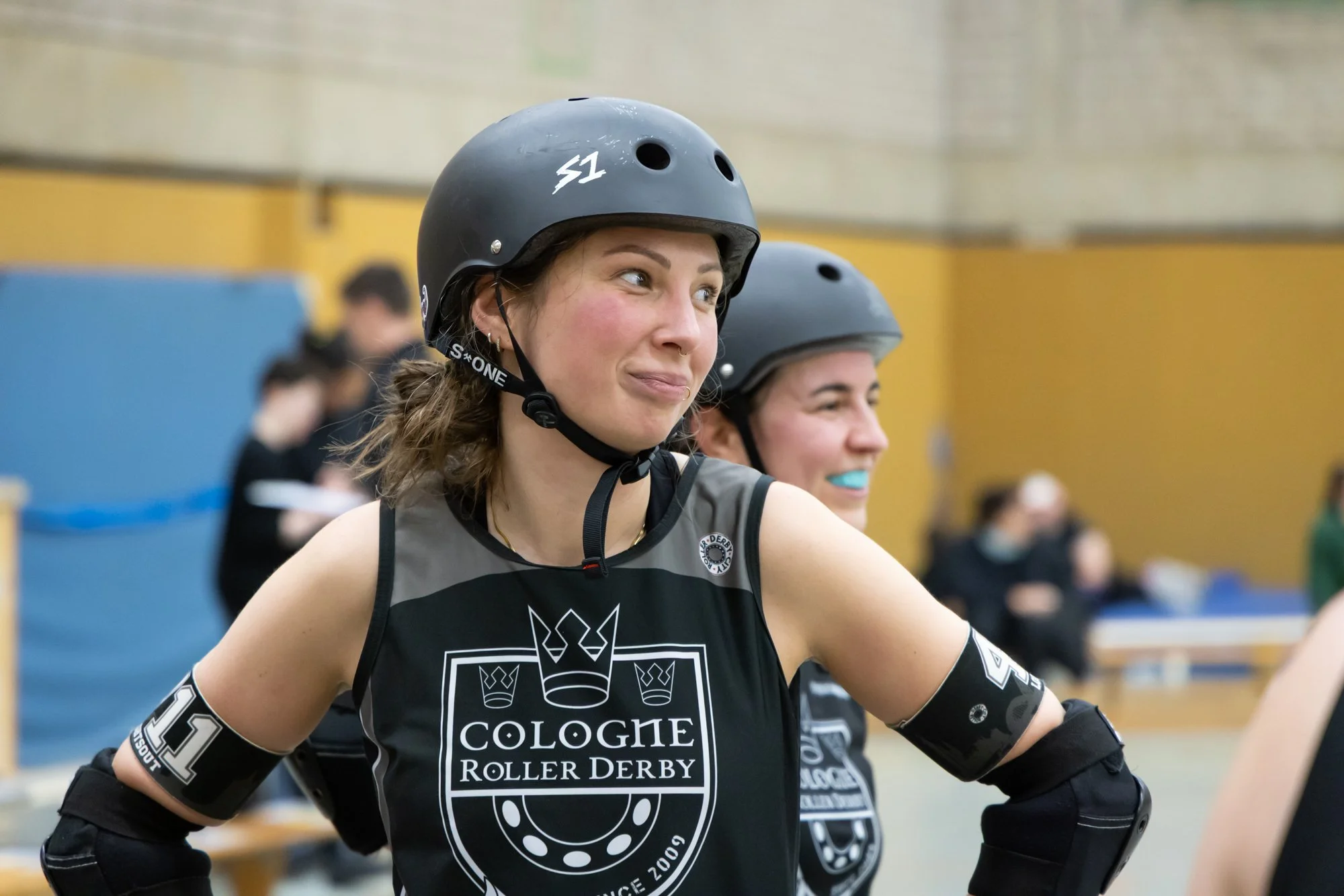 Zwei weibliche Rollschuhfahrerinnen tragen Helm und Trikots, während sie sich beim Cologne Roller Derby treffen, im Innenraum einer Turnhalle.