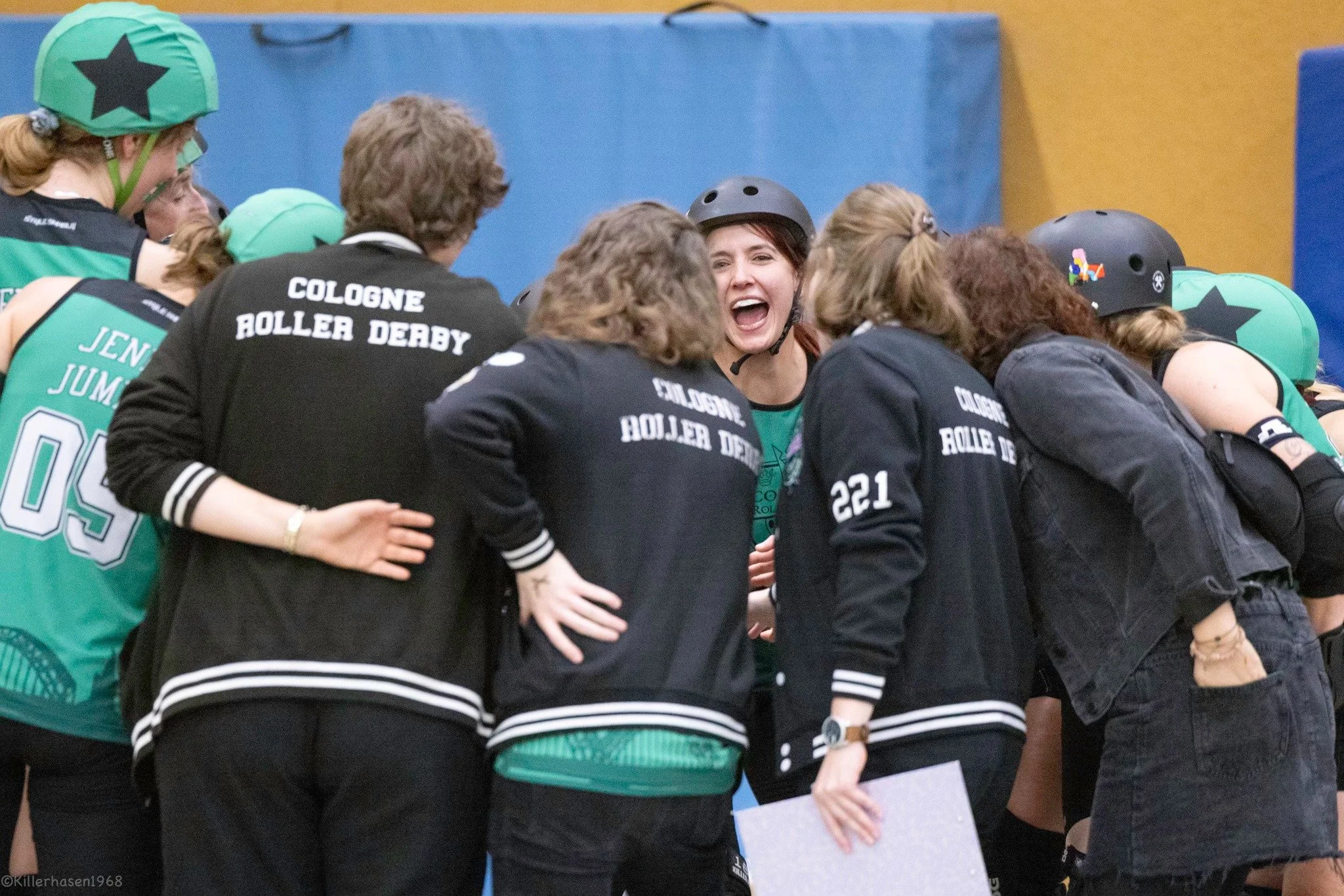 Gruppe von Frauen im Rollerderby-Team, die sich in einer Huddle versammelt und lächelnd sprechen, einige tragen Helme und Jacken mit Aufnähern und Teamnamen.