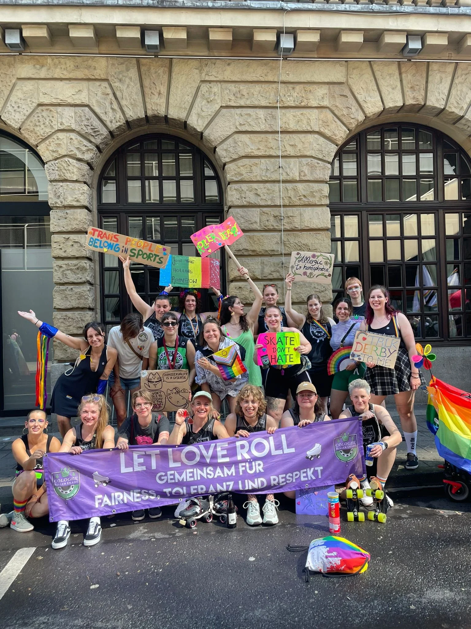 Gruppe von Menschen beim Pride-Parade, die einen Banner mit der Aufschrift 'LET LOVE ROLL GEMEINSAM FÜR FAIRNESS, TOLERANZ UND RESPEKT' halten, viele in Regenbogenfarben gekleidet, einige mit Rollschuhen, und sie tragen bunte Signale und Plakate für LGBTQ+-Rechte und Akzeptanz.
