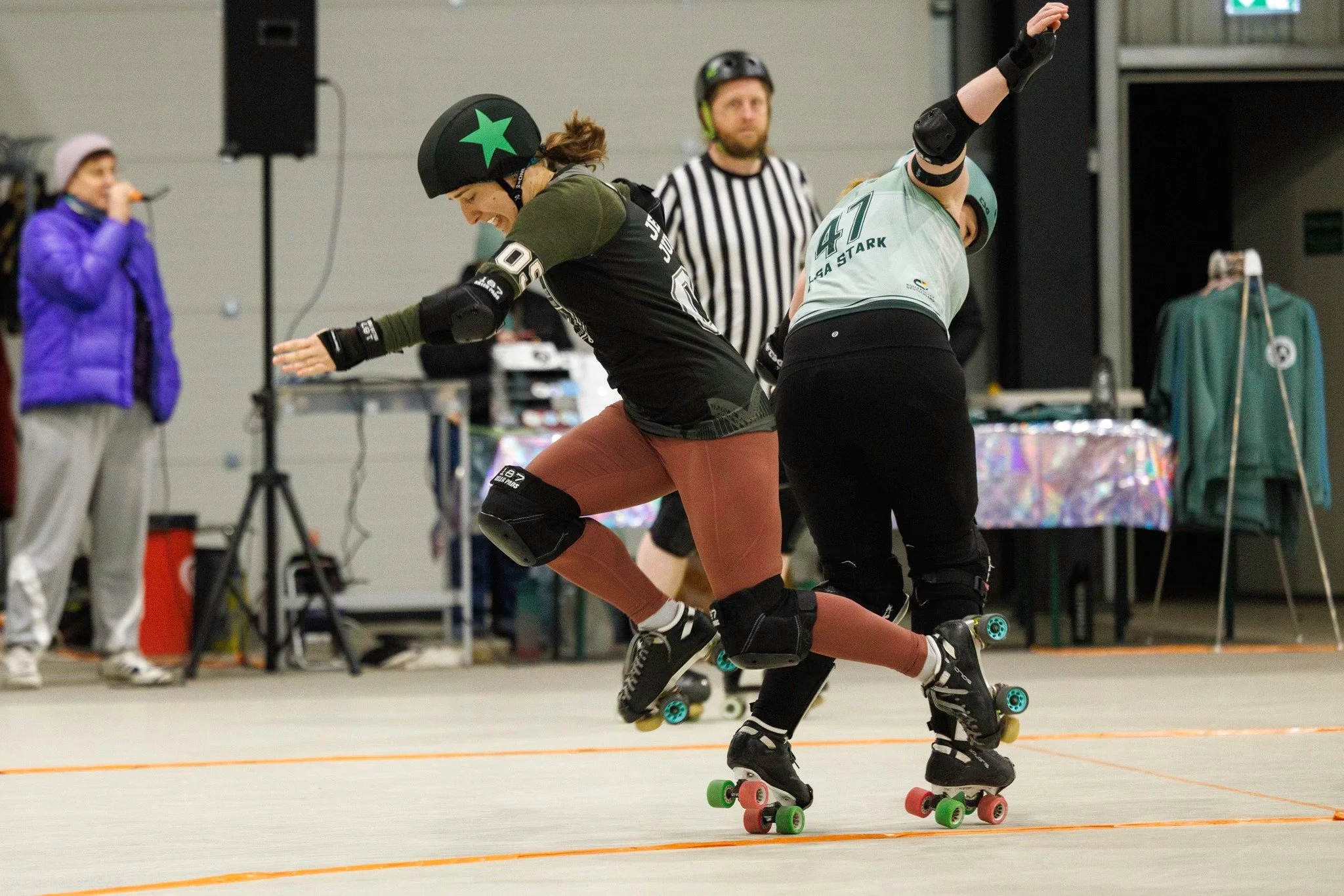 Zwei Rollschuhfahrer in Schutzkleidung beim Sturz auf der Rollschuhbahn, während ein Schiedsrichter und Zuschauer im Hintergrund zuschauen.