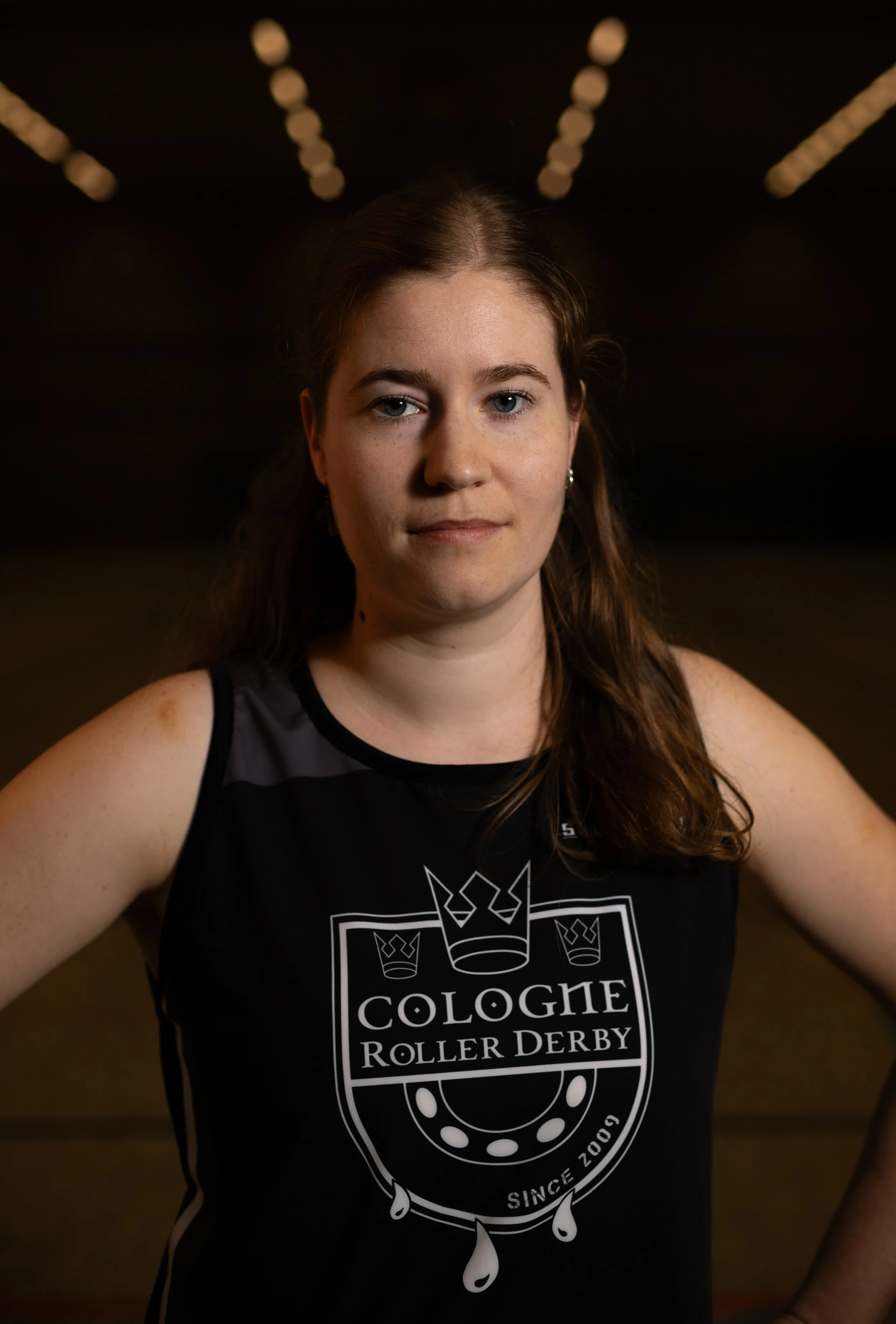 Junge Frau in einem schwarzen Trikot mit dem Logo 'Cologne Roller Derby', stehend im Rollschuhpark bei Nacht mit Lichtern im Hintergrund.