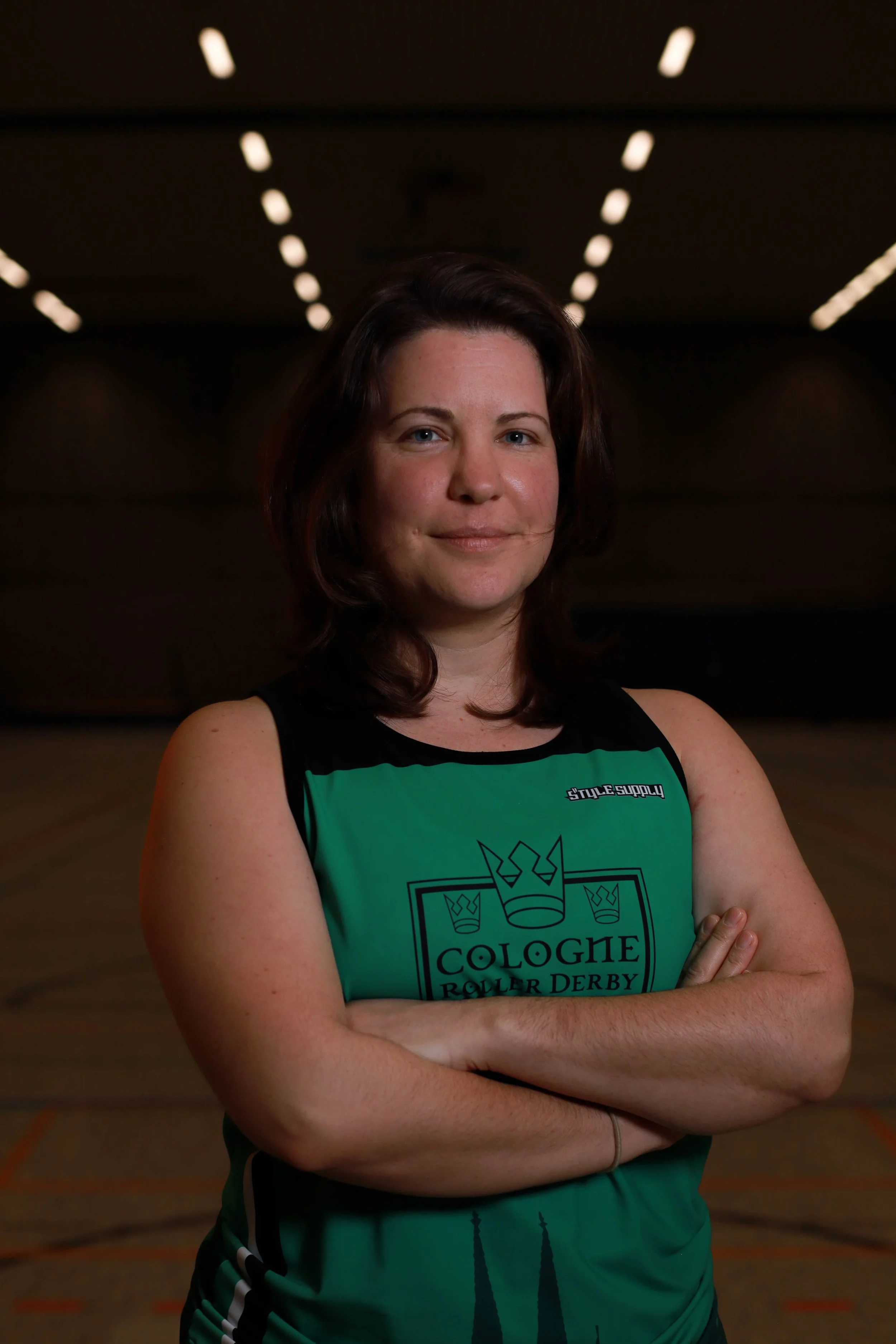 Frau in grün-schwarzem Trikot mit Armen verschränkt im Sporthalle, Kopf leicht nach rechts, lächelnd.