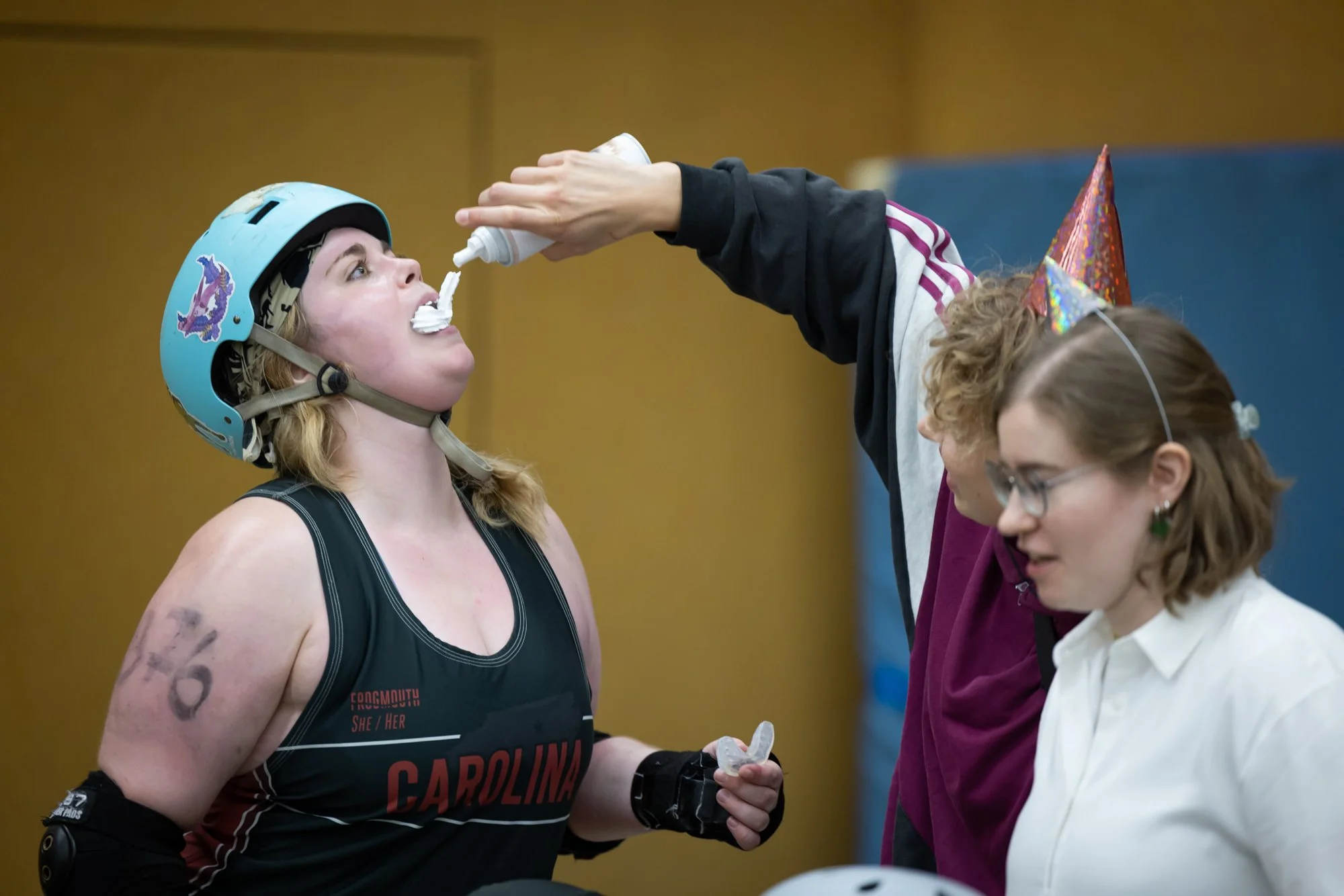 Eine Frau beim Rollschuhrennen bei einer Feier wird während eines Wettkampfs mit Schlagsahne beworfen, am Kopf trägt sie einen blauen Helm, die andere Person mit Partyhut spritzt Sahne auf sie, während eine weitere Person daneben zuschaut.