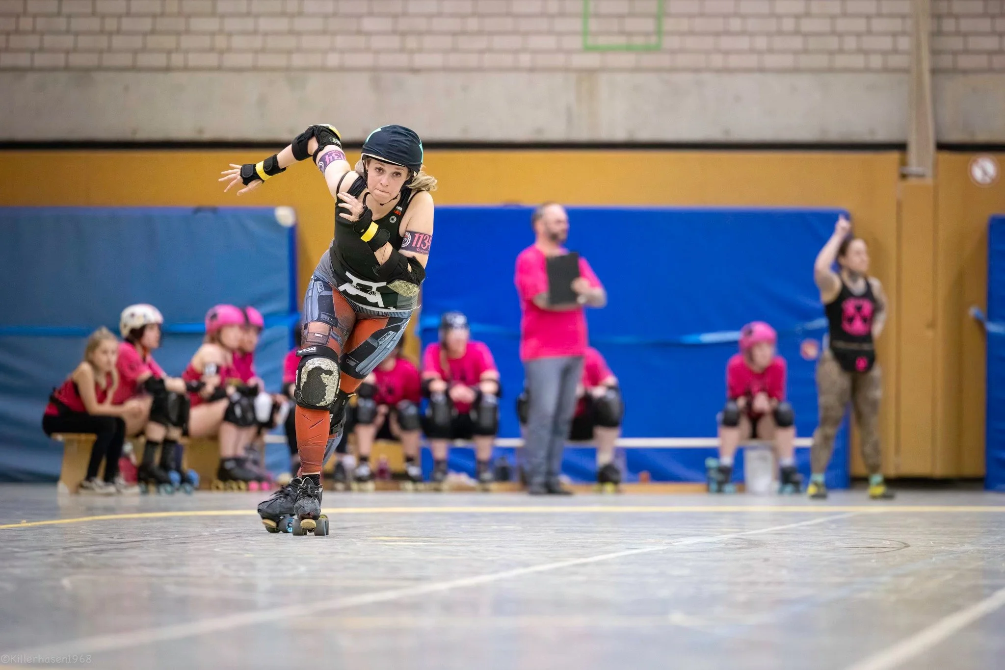 Skaterin beim Rollschuhlaufen in einer Turnhalle, mit Zuschauern und Trainern im Hintergrund, alle sind in Schutzkleidung.