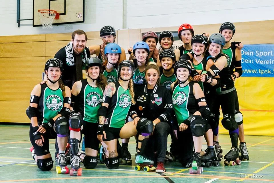 Eine Gruppe von weiblichen Rollschuhläuferinnen mit Schutzhelmen und Schutzausrüstung, zusammen mit einem Mann im Sporttrainer-Team, posieren in einer Turnhalle nach dem Rollenlauf. Die Frauen tragen grüne Trikots mit dem Logo 'Graveyard'.