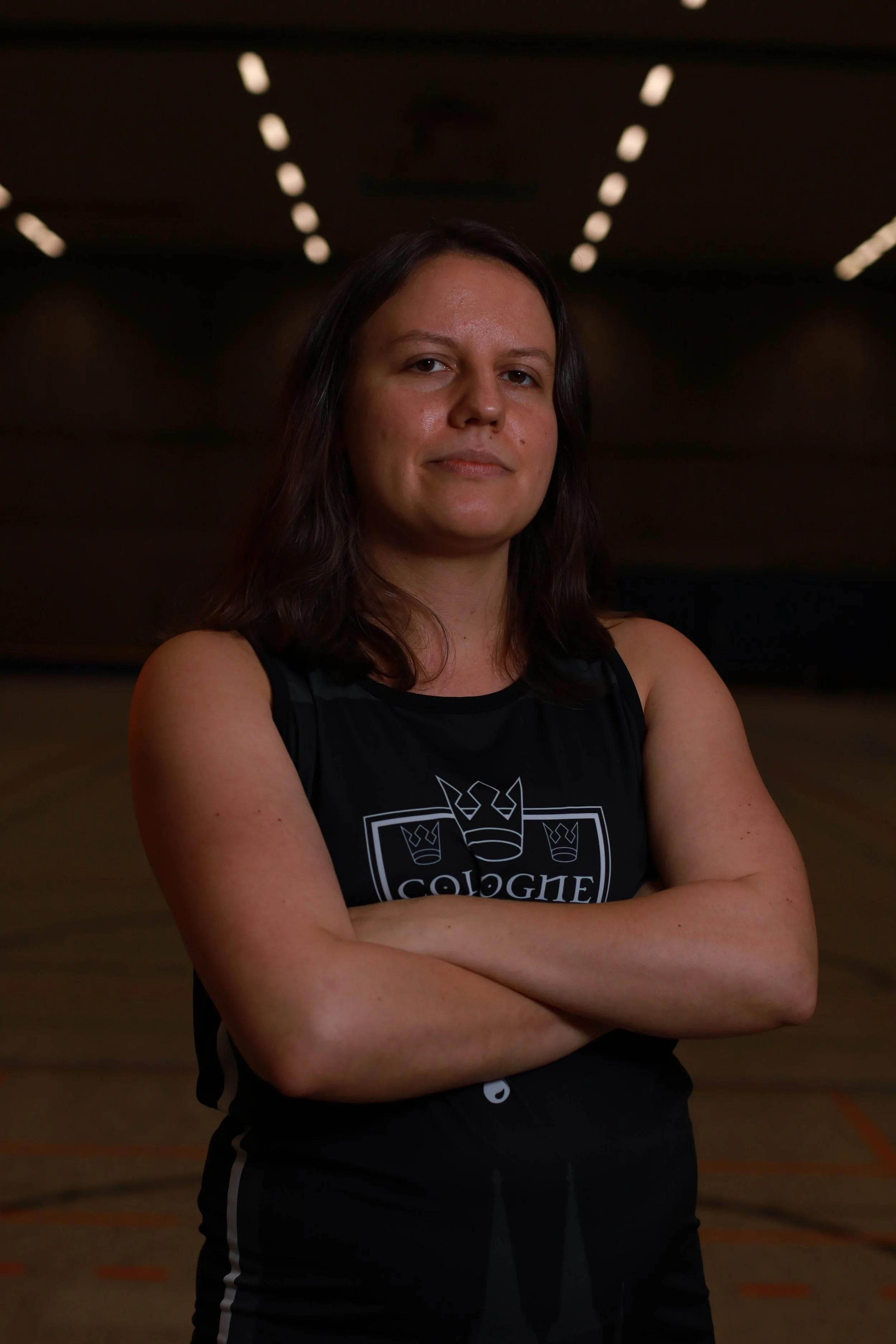 Eine Frau in Sportbekleidung steht in einer Turnhalle mit verschränkten Armen.