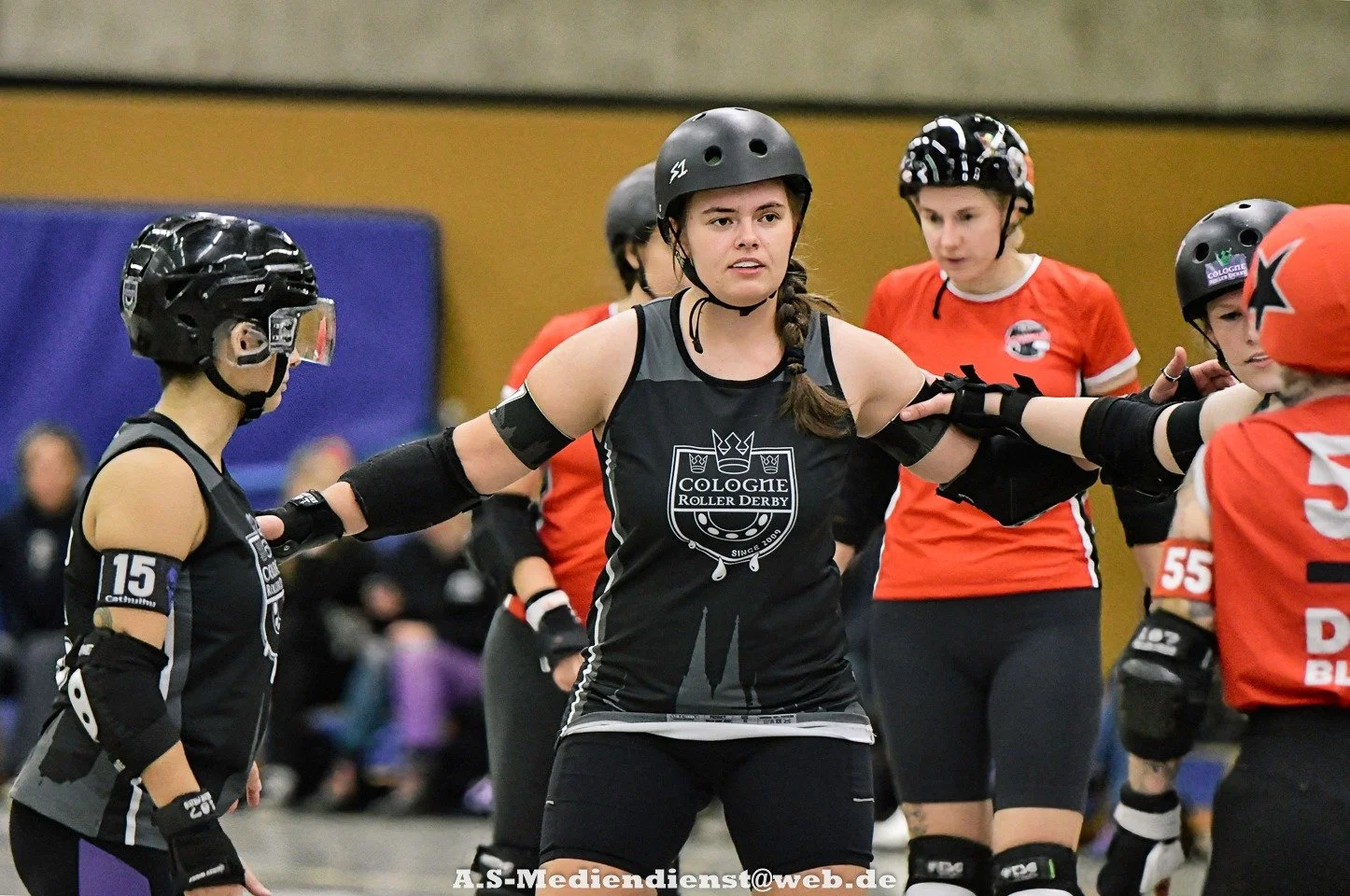 Skaterinnen beim Rollerderby auf der Rennbahn, eine Frau in der Mitte trägt ein schwarzes Trikot mit dem Logo "Cologne Roller Derby" und Helme, trägt Schutzausrüstung.