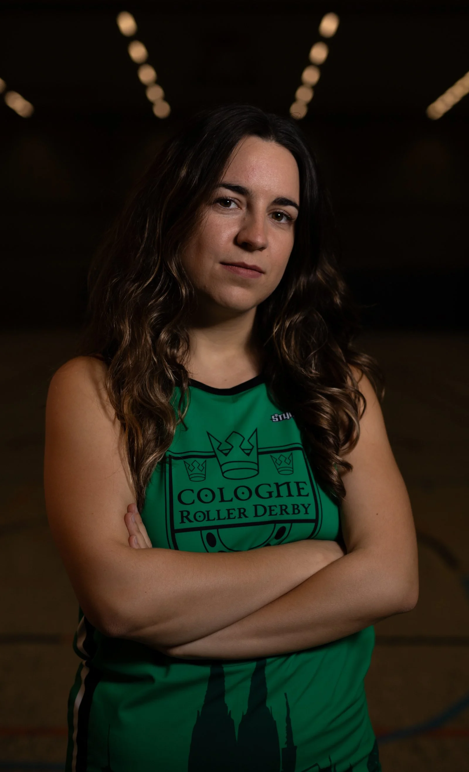 Junge Frau in grüner Sportkleidung mit 'Cologne Roller Derby' Logo, Arme verschränkt, in einer Hallenhalle bei Rollerderby-Event.