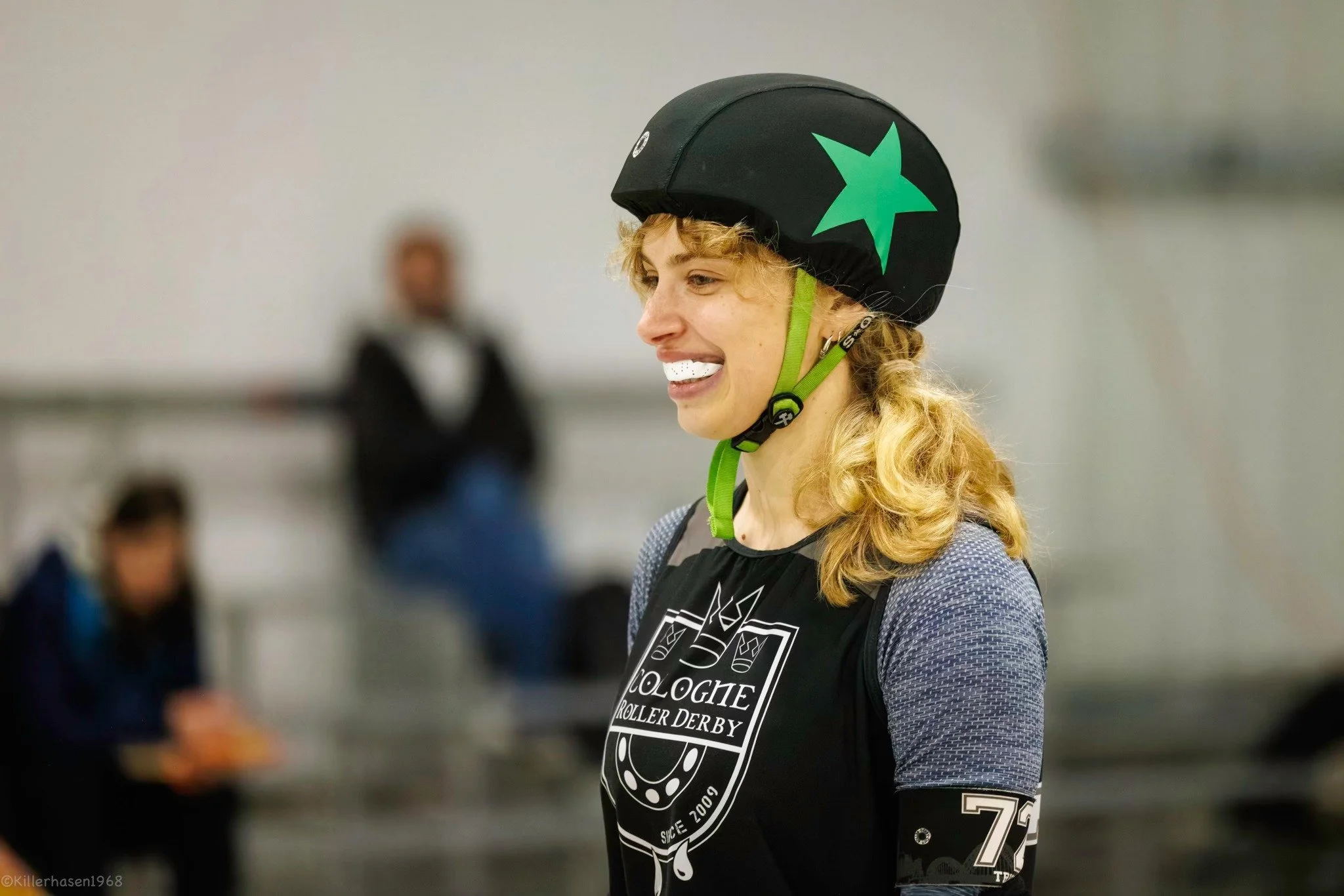 Eine junge Frau mit Locken trägt einen schwarzen Helm mit grüner Sternmarkierung und eine Schutzbrille. Sie lächelt bei einer Rollschuh-Veranstaltung im Indoor-Sportzentrum.