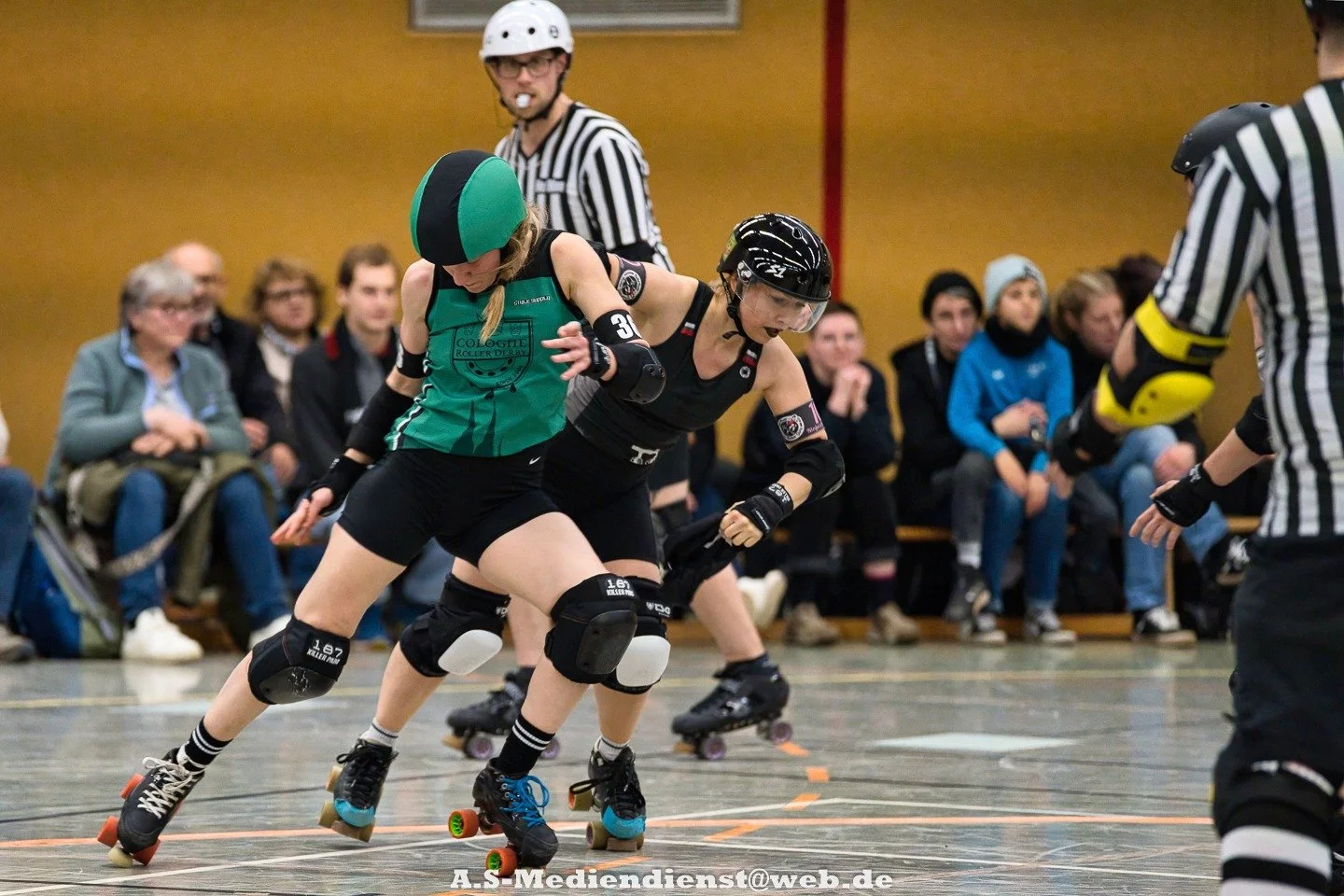 Zwei Rollschuhfahrerinnen beim Rollschuhkampf, eine in grün, die andere in schwarz, kämpfen in einem Indoor-Sportzentrum, während mehrere Kampfrichter und Zuschauer zuschauen.