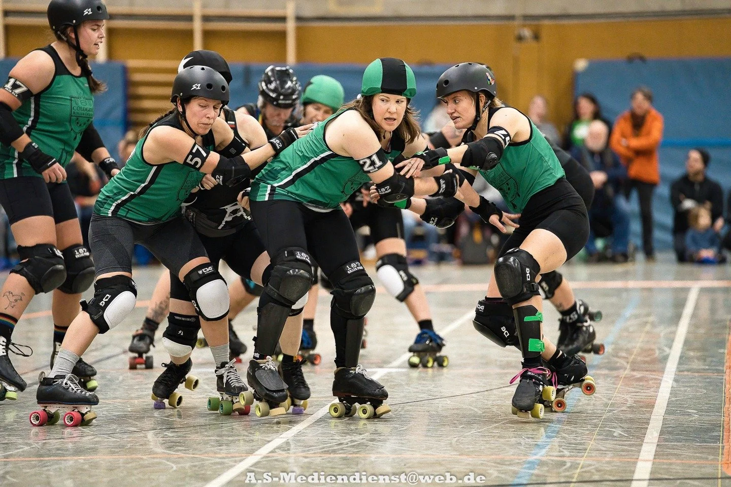 Roller Derby Frauen in Teamuniformen beim Spiel auf der Rollschuhbahn, in Aktion, mit Schutzausrüstung, im Hintergrund Zuschauer.