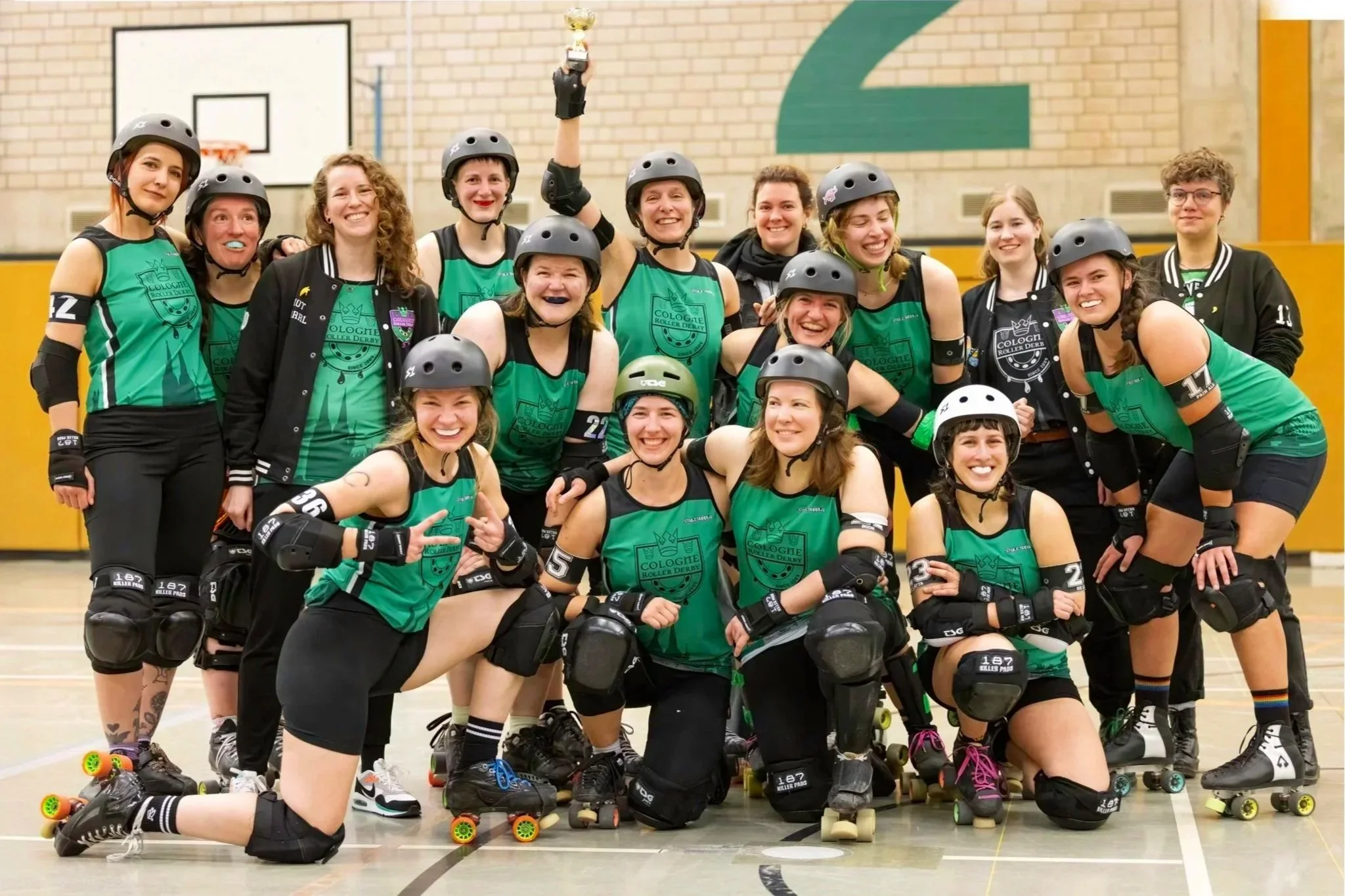 Gruppe von Vertretern des Rollerderbys im Sportlertrikot mit Helmen und Schutzausrüstung, die im Basketball-Stadion feiern und einen Pokal hochhalten.