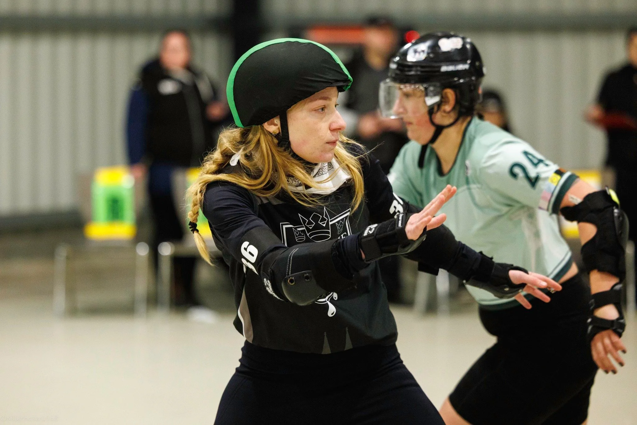 Zwei Rollschuhfahrerinnen trainieren in einer Halle, eine trägt einen schwarzen Helm und schwarze Kleidung, die andere einen schwarzen Helm mit klarem Gesichtsschutz und grünes Trikot.
