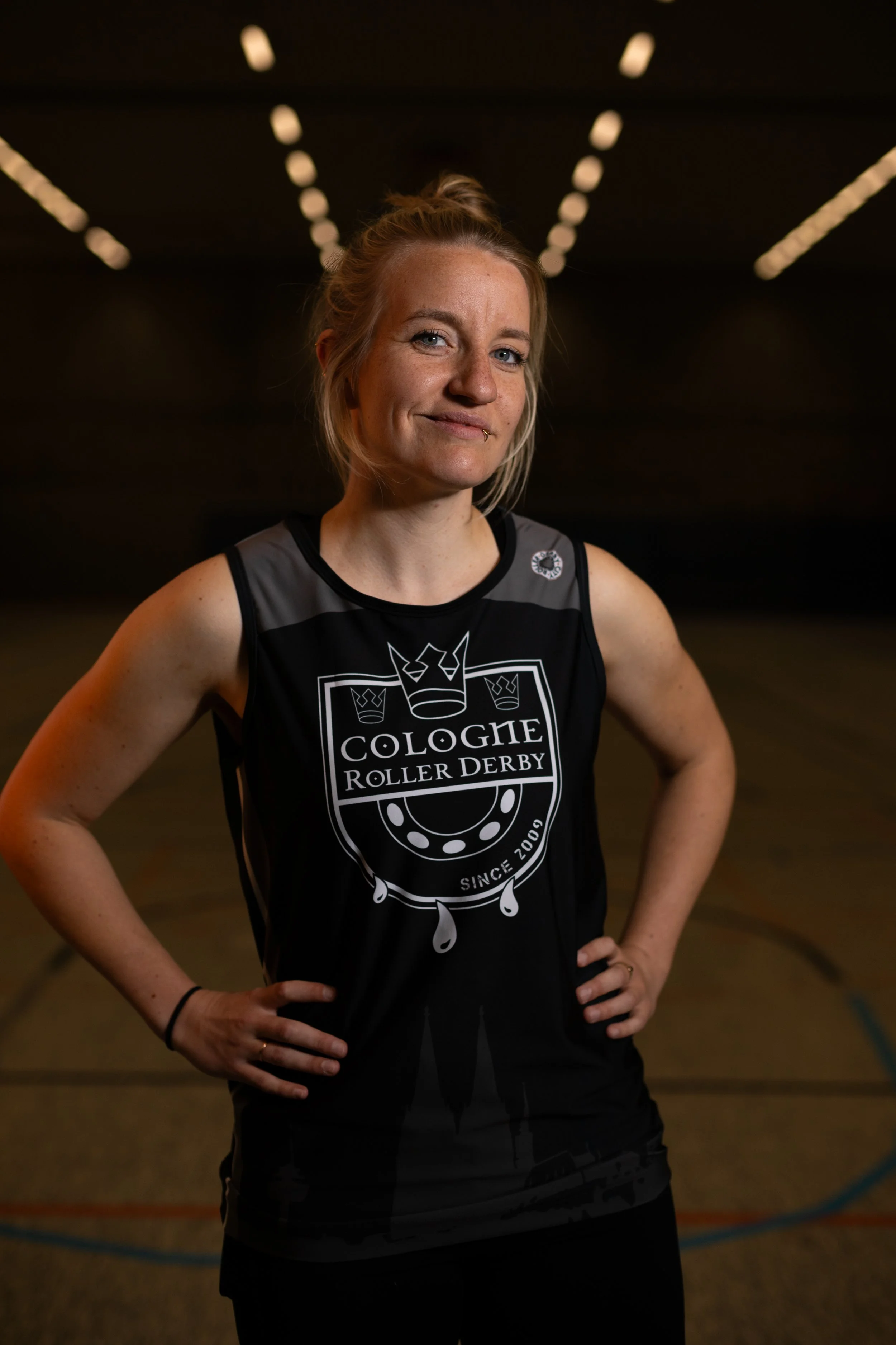 Eine Frau trägt ein schwarzes Trikot mit dem Logo "Cologne Roller Derby" auf einer Rollschuhbahn.
