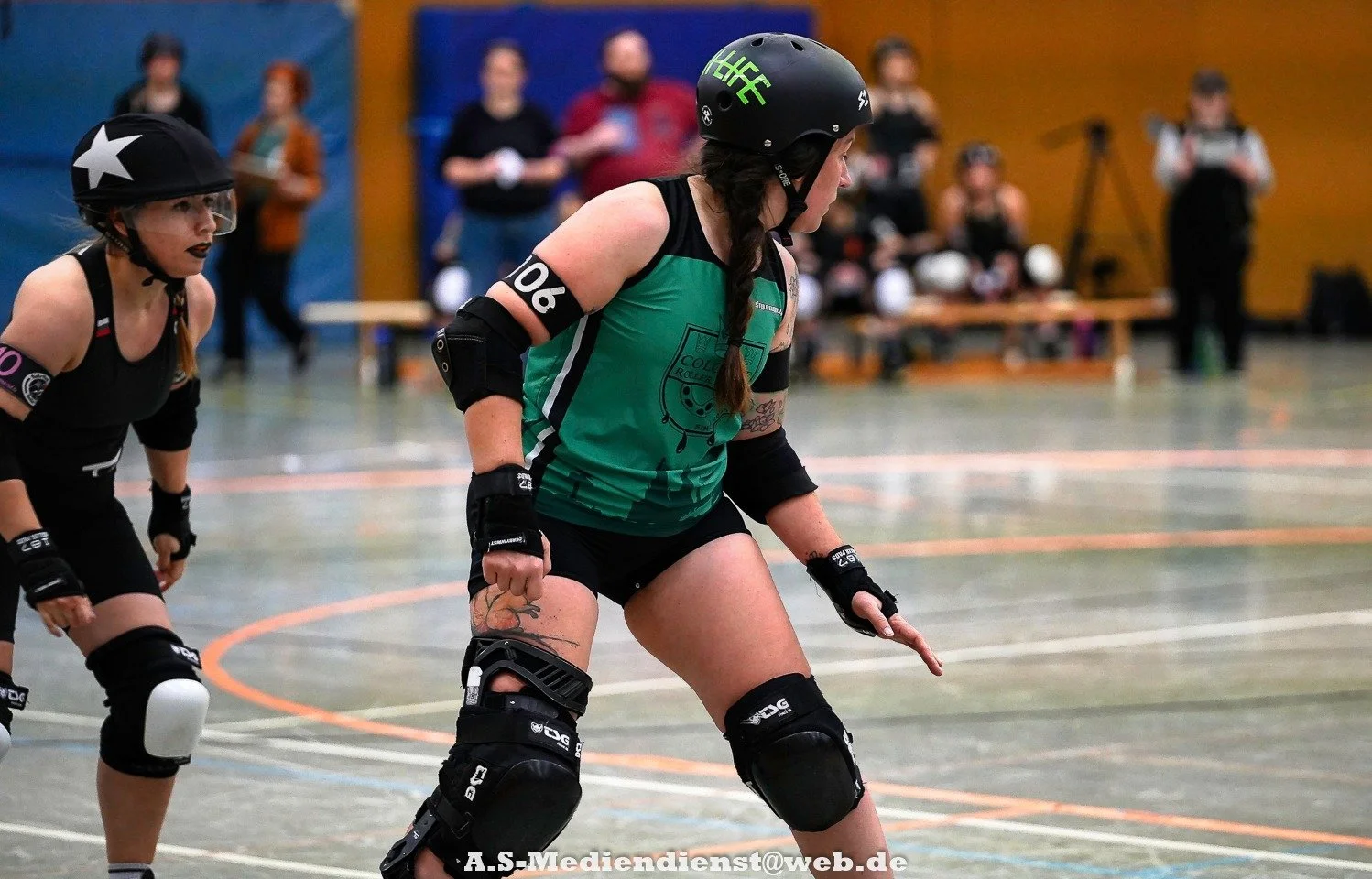 Roller Derby Spielerinnen in Aktion auf der Bahn, tragen Helme, Schutzkleidung und Trikots, in einem Hallenstadion mit Zuschauern im Hintergrund.
