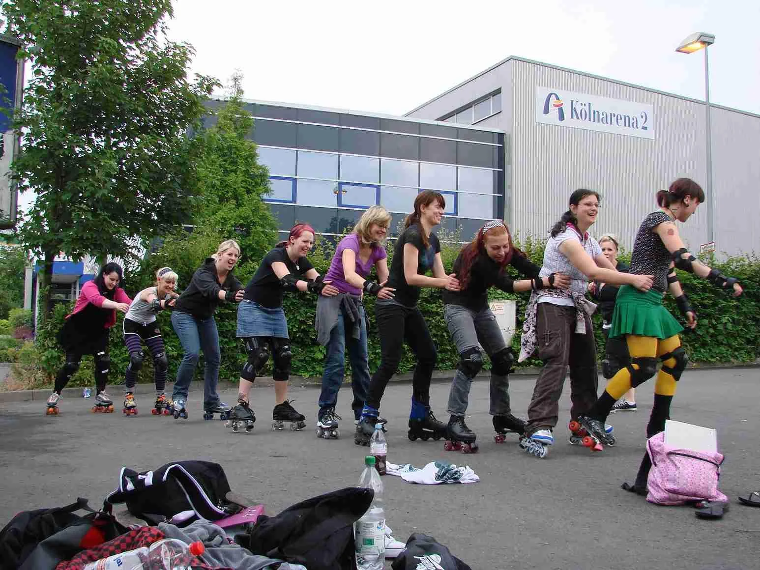 Eine Gruppe von Frauen rollschuhfahrend in einer Linie draußen vor einem Gebäude mit der Aufschrift 'Kölnerarena2'.