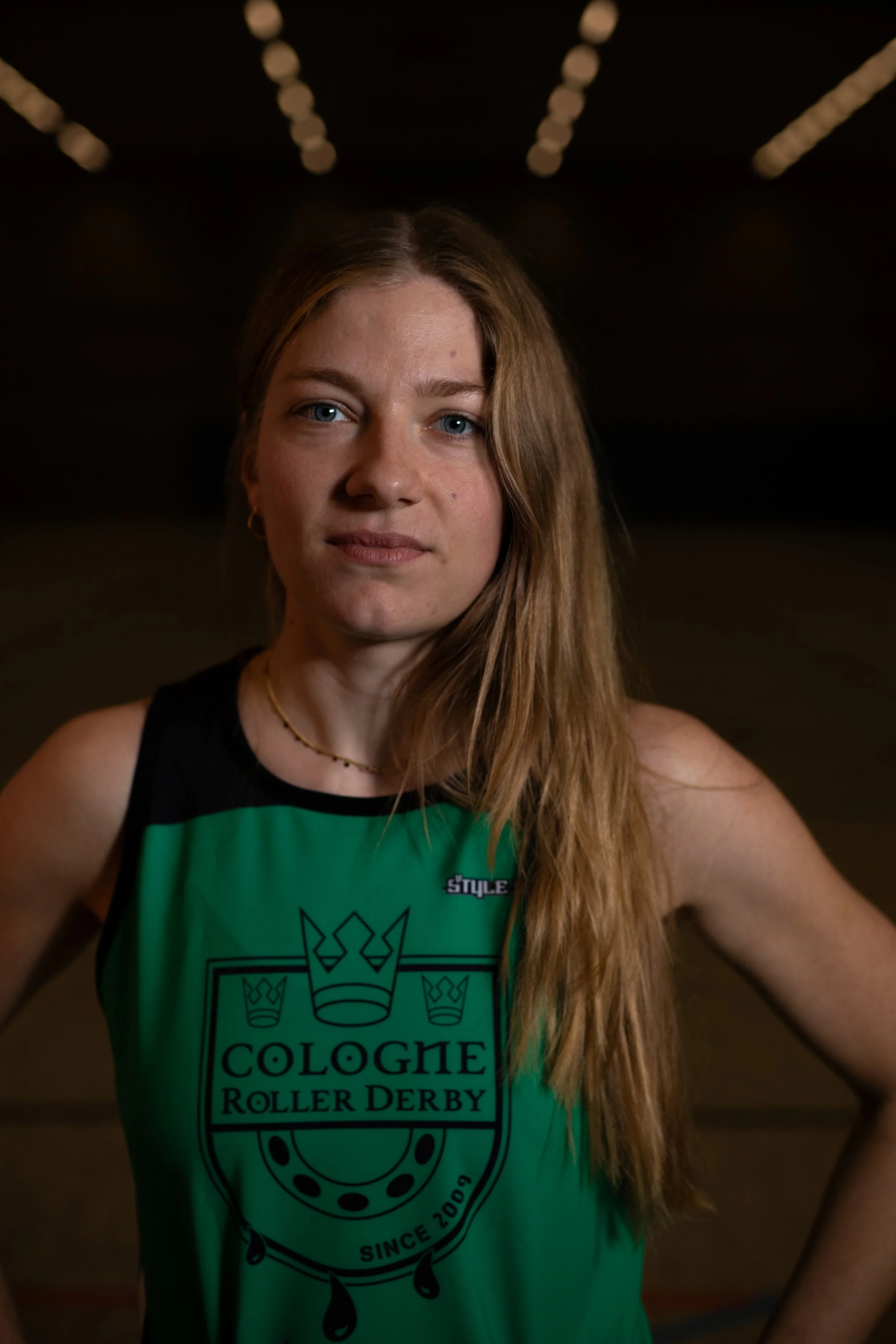 Junge Frau mit langen, light-braunen Haaren trägt ein grünes Sportshirt mit dem Logo "Cologne Roller Derby" und posiert in einer dunklen, urbanen Umgebung.