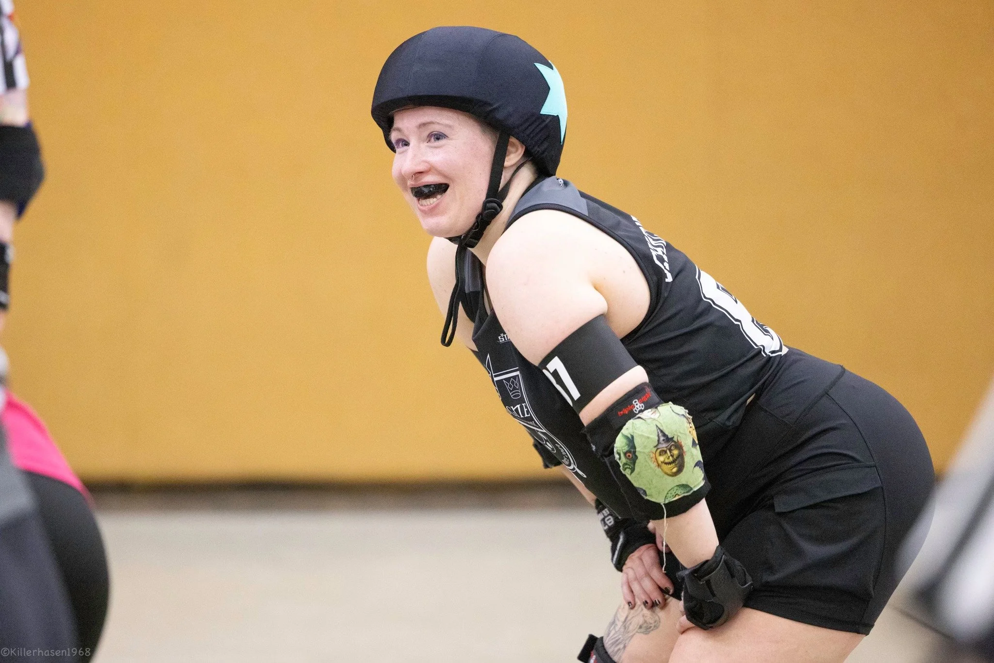 Eine Frau beim Rollerderby trägt Schutzhelm, Arm- und Ellbogenschoner, und lacht während des Spiels.