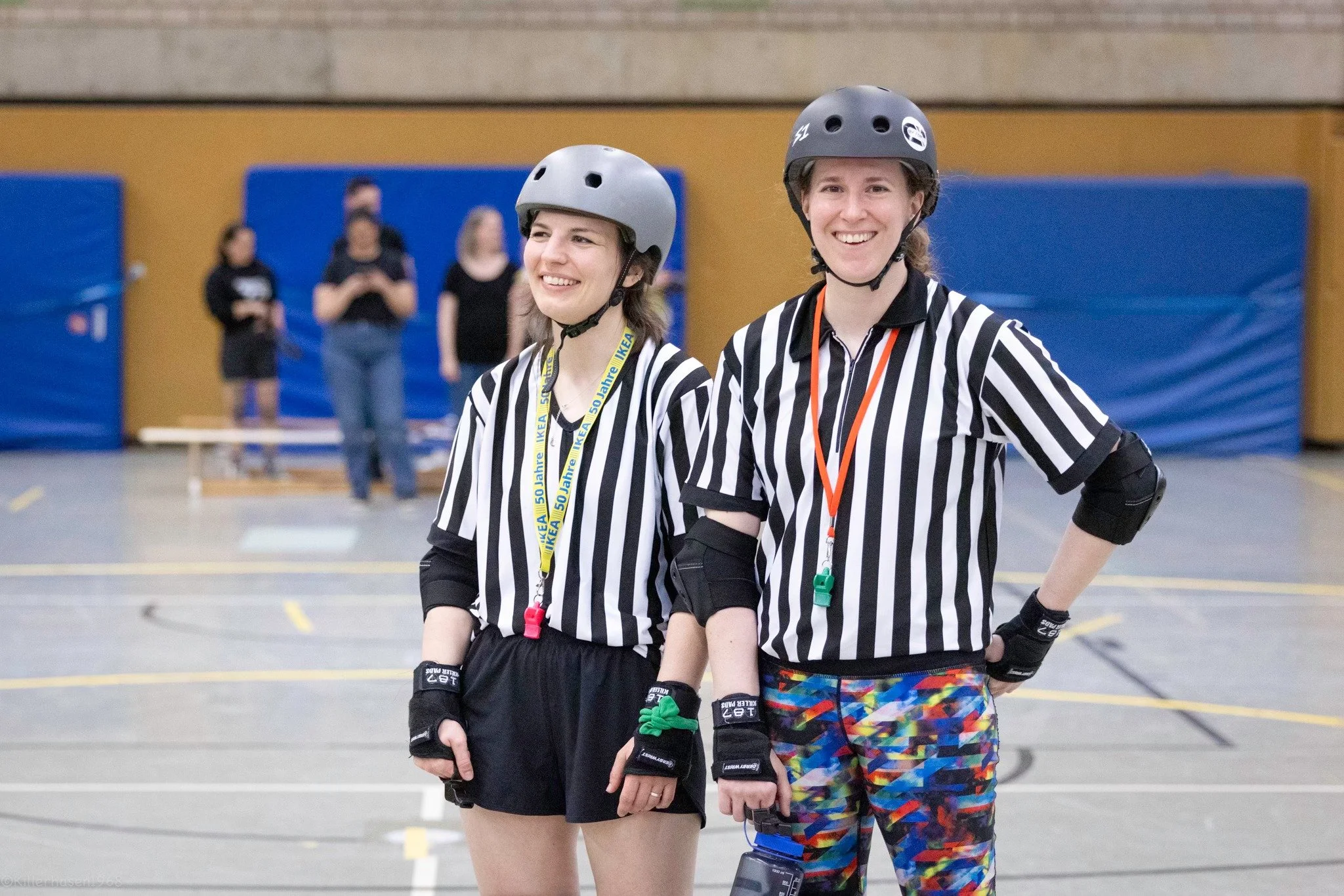 Zwei weibliche Roller Derby Schiedsrichterinnen in schwarz-weiß gestreiften Trikots, Helm und Schutzausrüstung, die in einer Turnhalle auf dem Spielfeld stehen und lächeln.