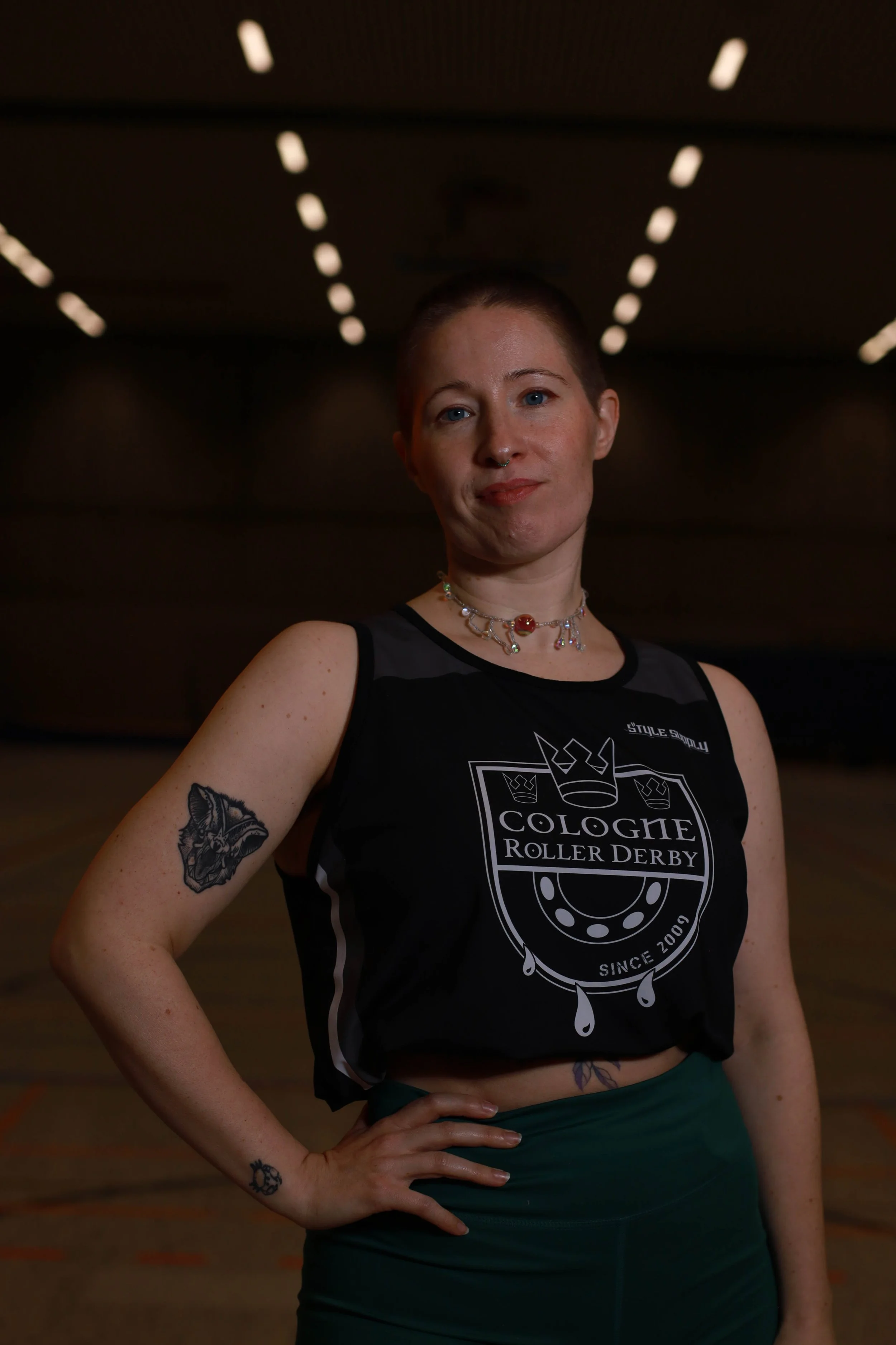 Eine junge Frau mit kurzen Haaren, Tattoos am Arm, trägt ein schwarzes Top mit dem Logo "COLOGNE ROLLER DERBY" und steht in einer Turnhalle mit dunklem Hintergrund.